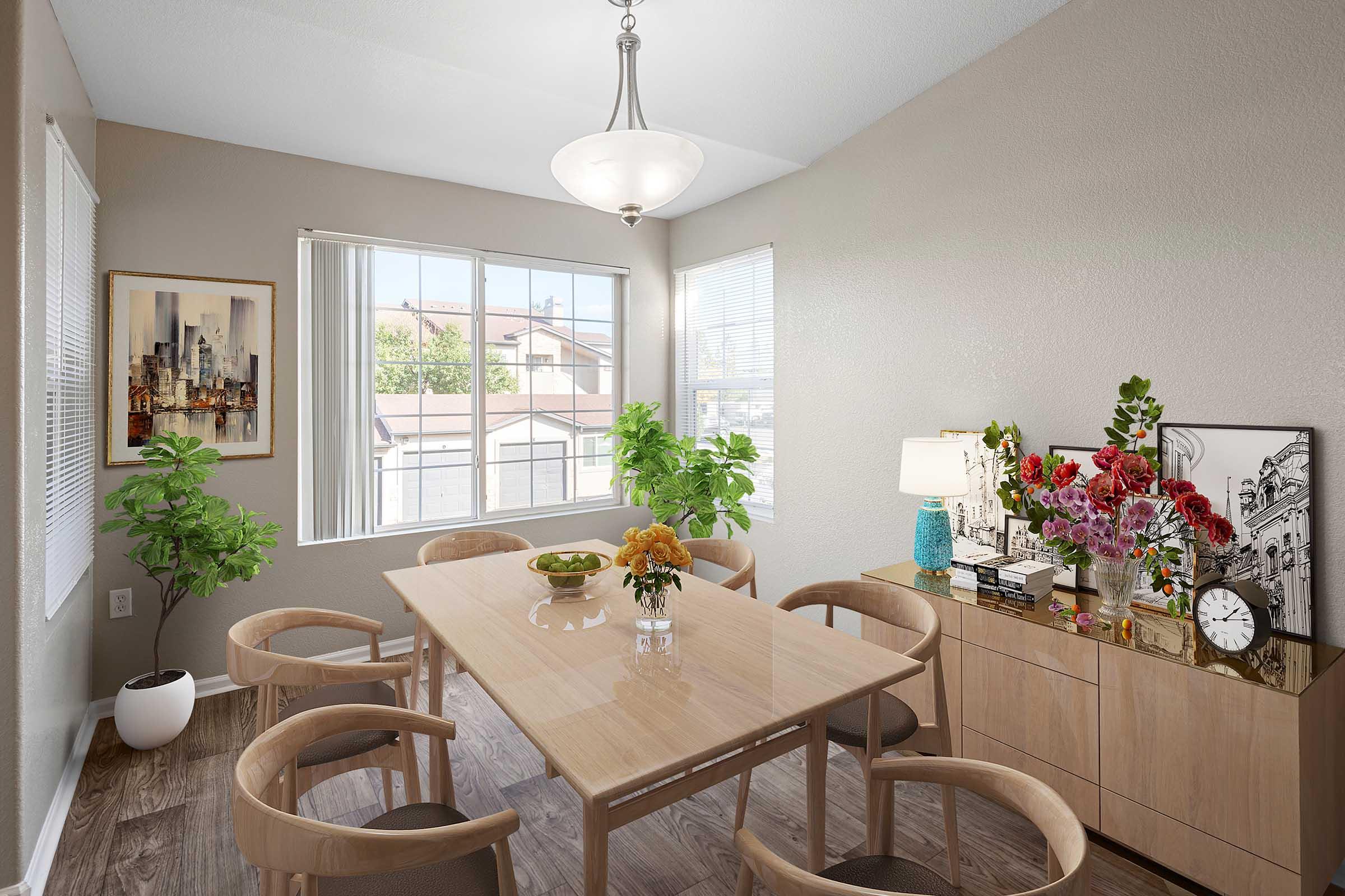 A well-lit dining room featuring a wooden table surrounded by circular chairs. A vase of flowers sits on the table, and a sideboard displays additional flowers, books, and a clock. Large windows allow natural light to illuminate the room, and artwork depicting a city skyline is hung on the wall.