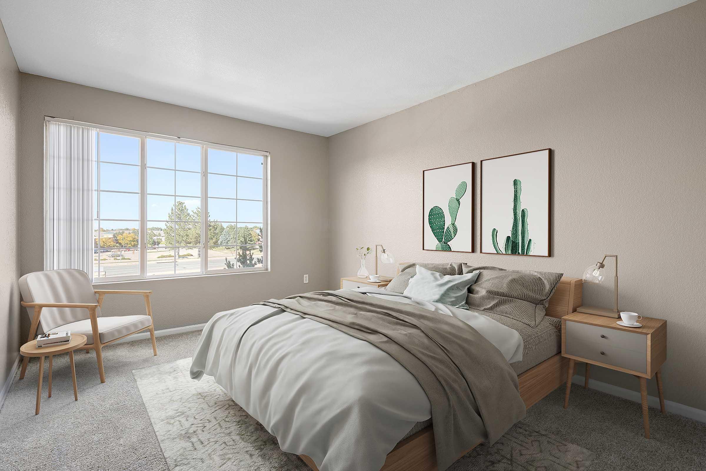 A modern bedroom featuring a neatly made bed with soft gray bedding, a light wood nightstand and lamp, and a comfortable armchair in the corner. Two framed cactus prints adorn the wall, and large windows allow natural light to brighten the space, showcasing a view of greenery outside.