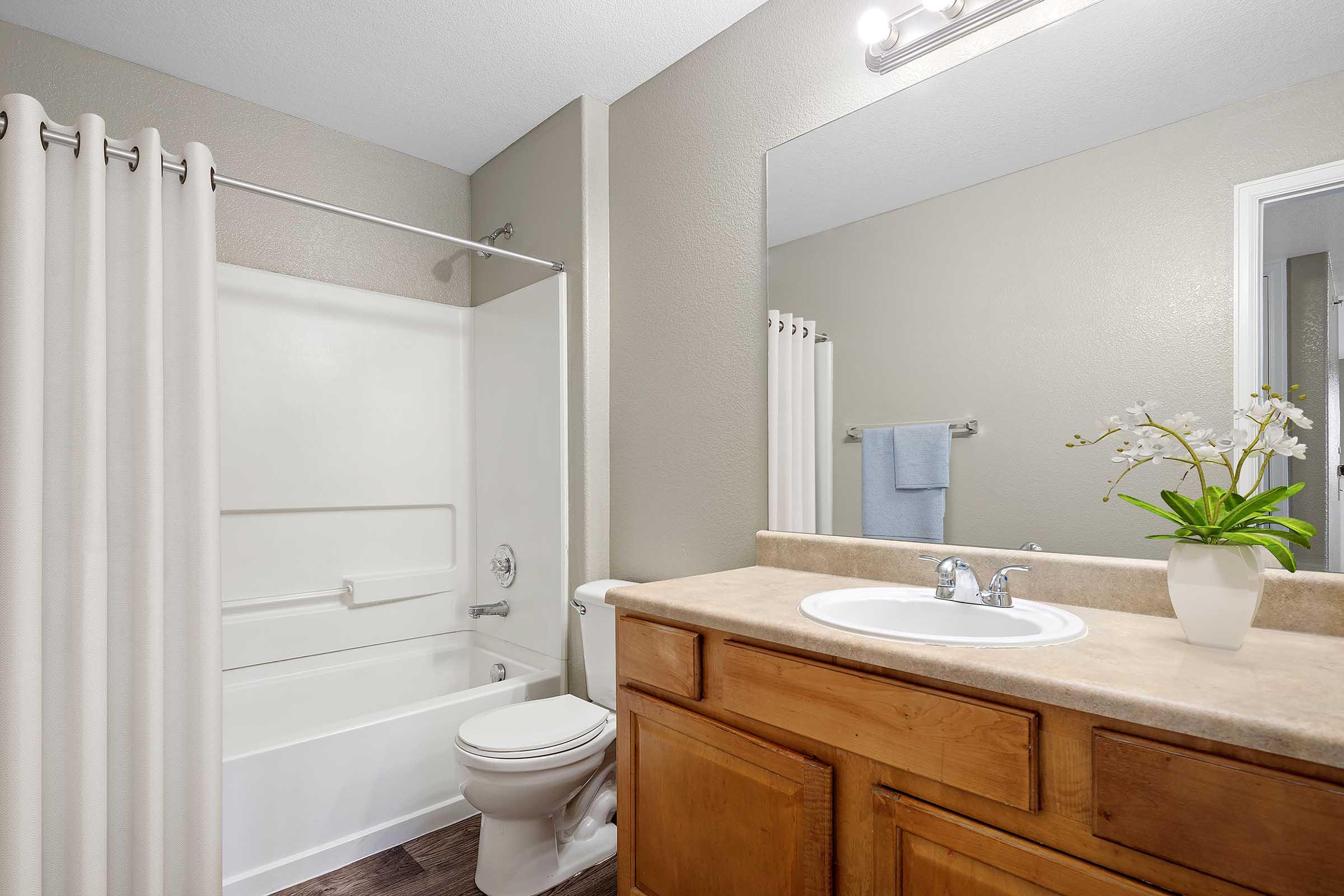A clean and modern bathroom featuring a white bathtub and shower combination, a wooden vanity with a sink, and a large mirror above. Neutral-colored walls and a towel rack with a blue towel complement the space. A potted plant adds a touch of greenery.