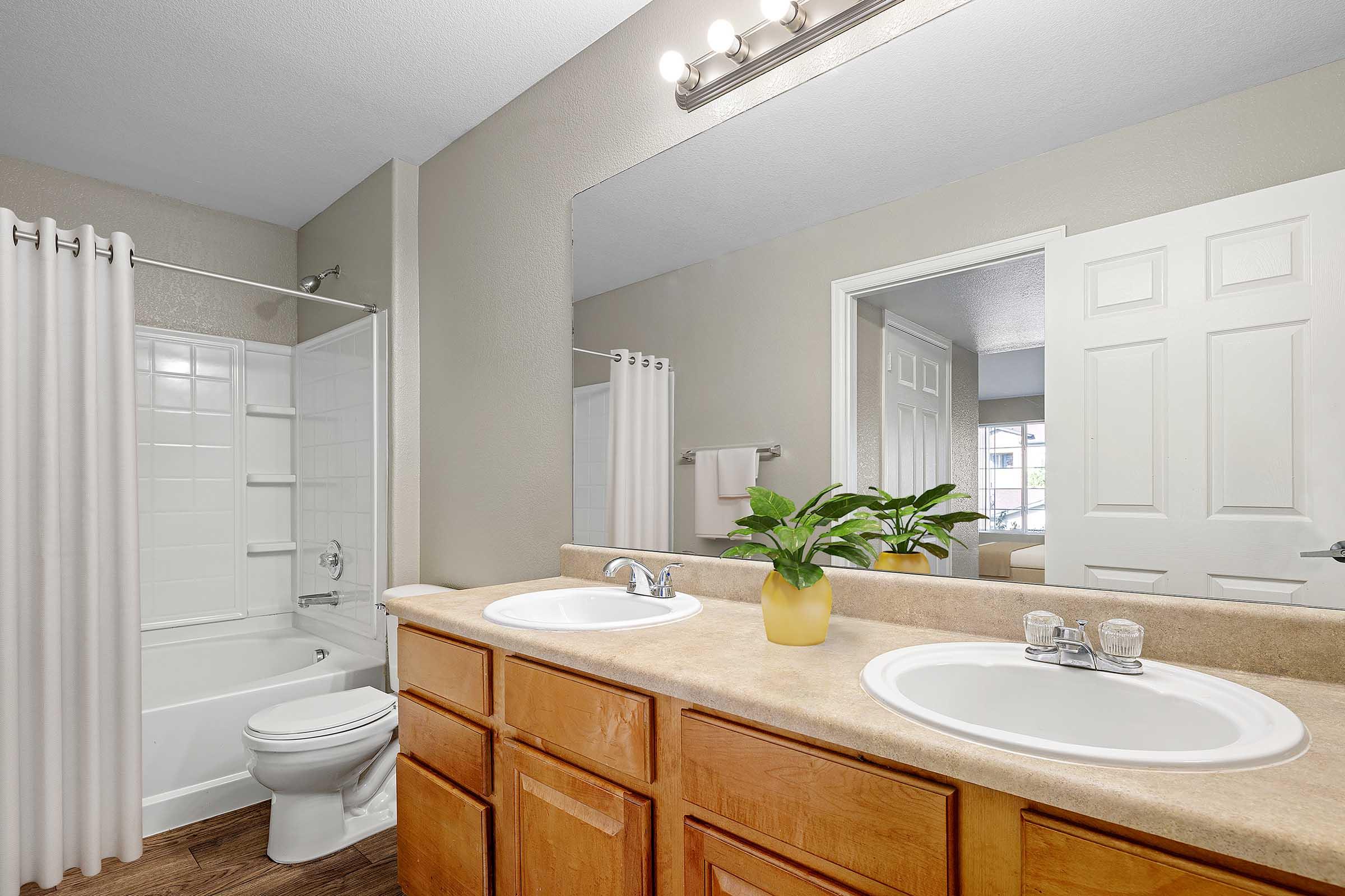 A modern bathroom featuring a double vanity with two sinks, wooden cabinetry, a large mirror, and a bathtub with a shower curtain. A potted plant adds a touch of greenery. The walls are painted in neutral tones, and there is a partially open door leading to another room.