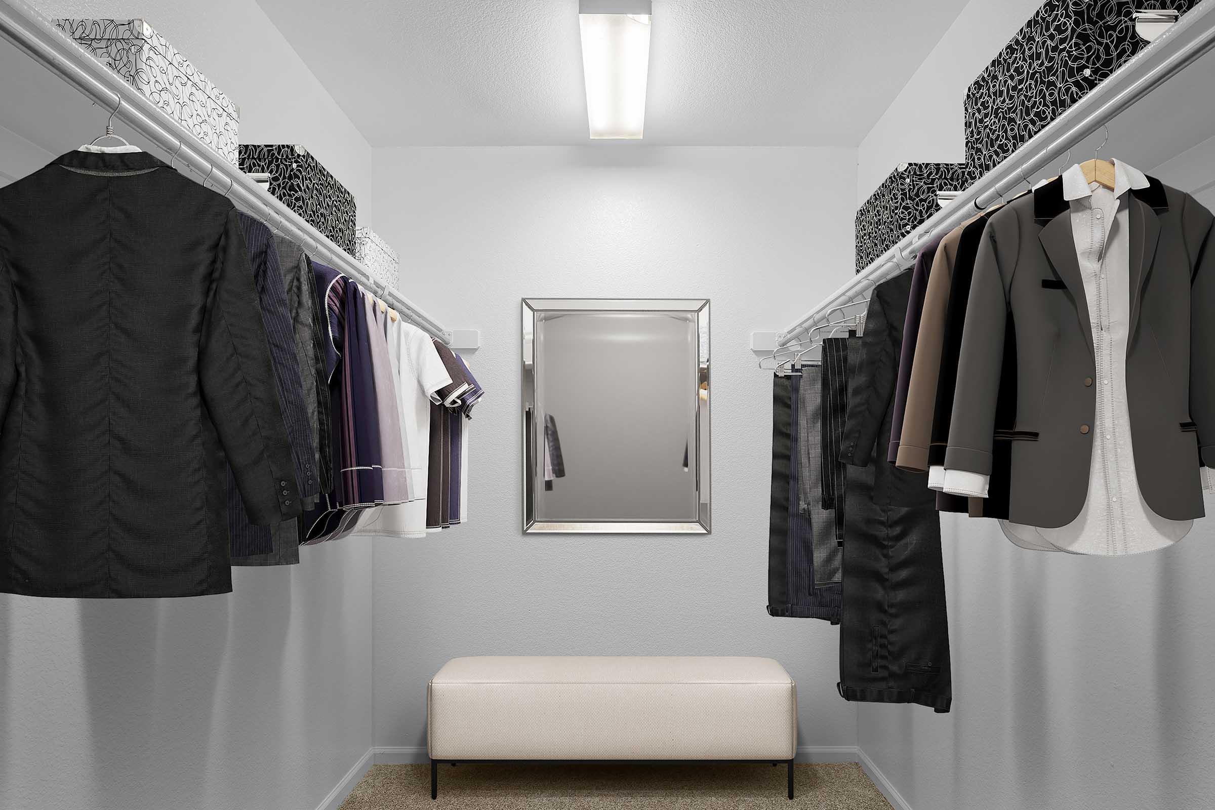 A well-organized walk-in closet featuring hanging suits, shirts, and accessories on either side. A full-length mirror is positioned on the wall across from a cream-colored bench in the center. The closet has a clean, modern design with gray walls and stylish storage boxes on the top shelves.