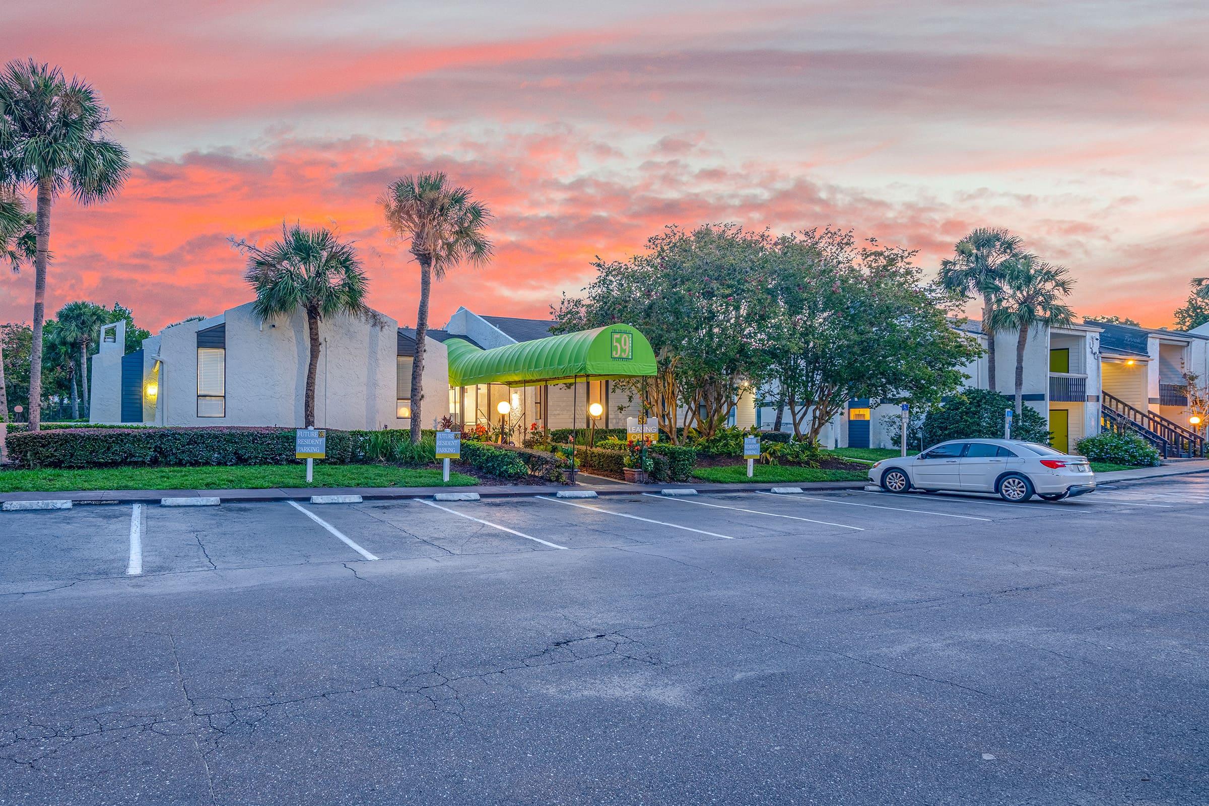 A parking lot in front of a residential building with a green canopy at sunset, featuring palm trees and colorful sky.