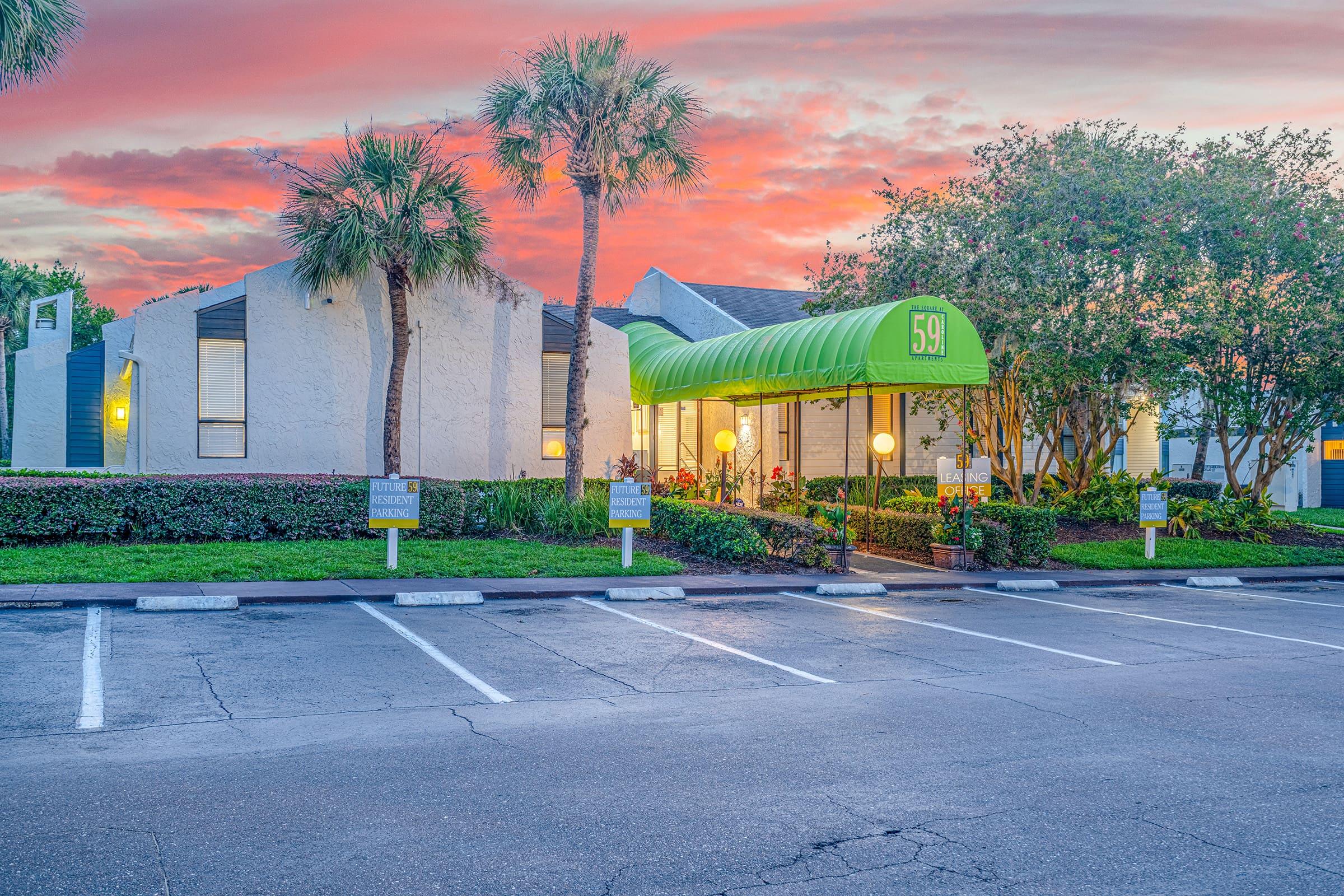 A building with a green awning displaying the number 59, surrounded by palm trees and vibrant landscaping, under a colorful sunset sky. Parking spaces are visible in the foreground.