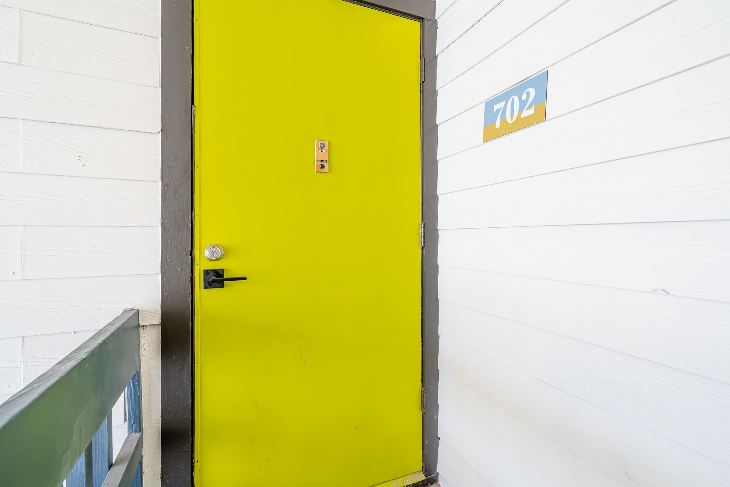 A bright yellow door with a small number 702 on a blue sign next to it. The door has a simple lock and is set against a backdrop of white wooden siding. A green railing is visible in the foreground, adding to the colorful and inviting appearance of the entrance.