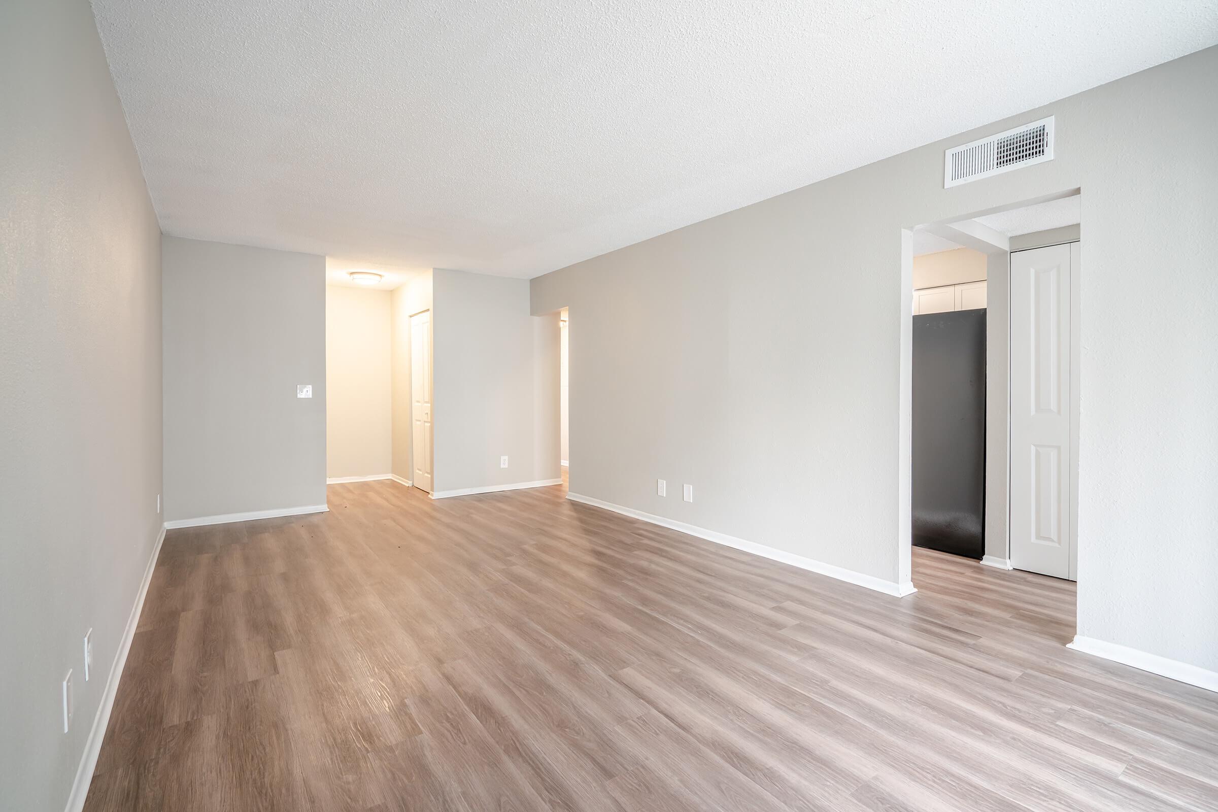 Spacious, empty living area showcasing light-colored walls and modern vinyl flooring. Two doorways lead to adjacent rooms, with ample natural light enhancing the open layout. The space is ready for furnishing, offering a neutral backdrop for personalization.