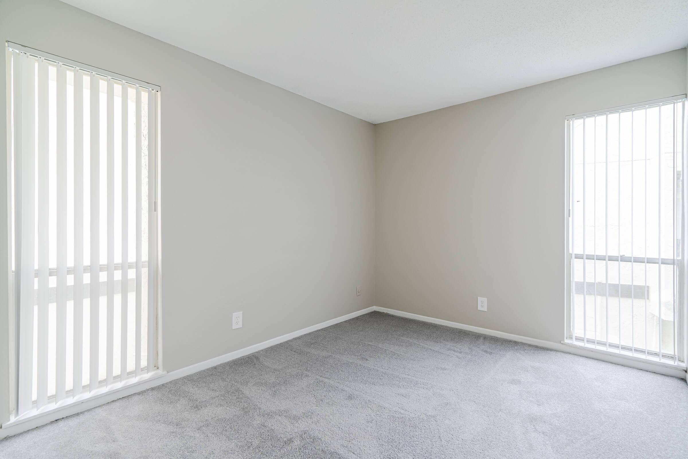 A vacant room with light gray carpet and neutral-colored walls. Two vertical-blind windows allow natural light to enter, creating a spacious and airy feeling. The room is empty, with no furniture or decorations, highlighting the potential for customization.