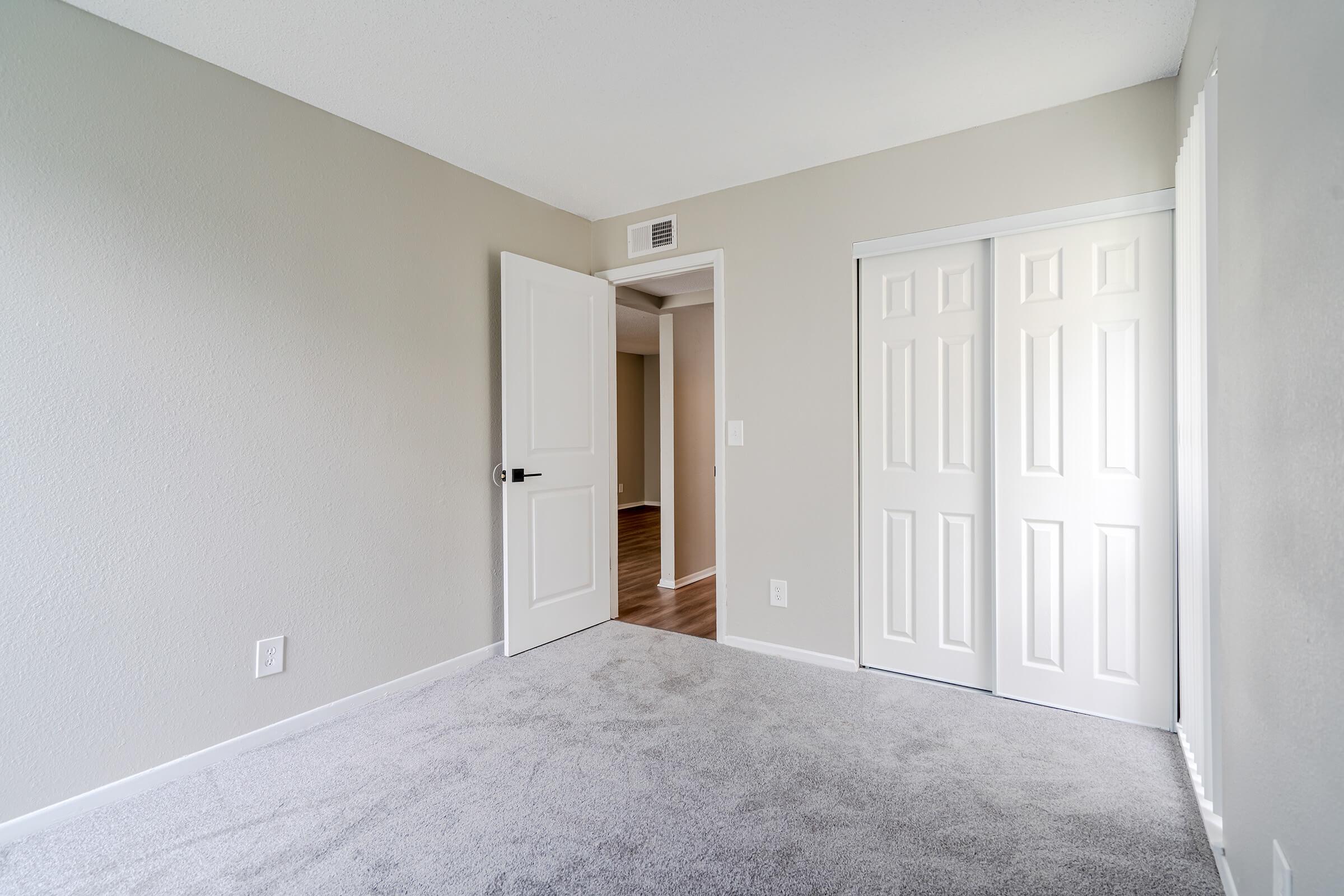 A cozy, empty room with light gray walls and soft carpet. One door leads to another area, while a closet with double doors is visible on the right. Natural light brightens the space, creating a clean and inviting atmosphere, perfect for personal decoration or use.