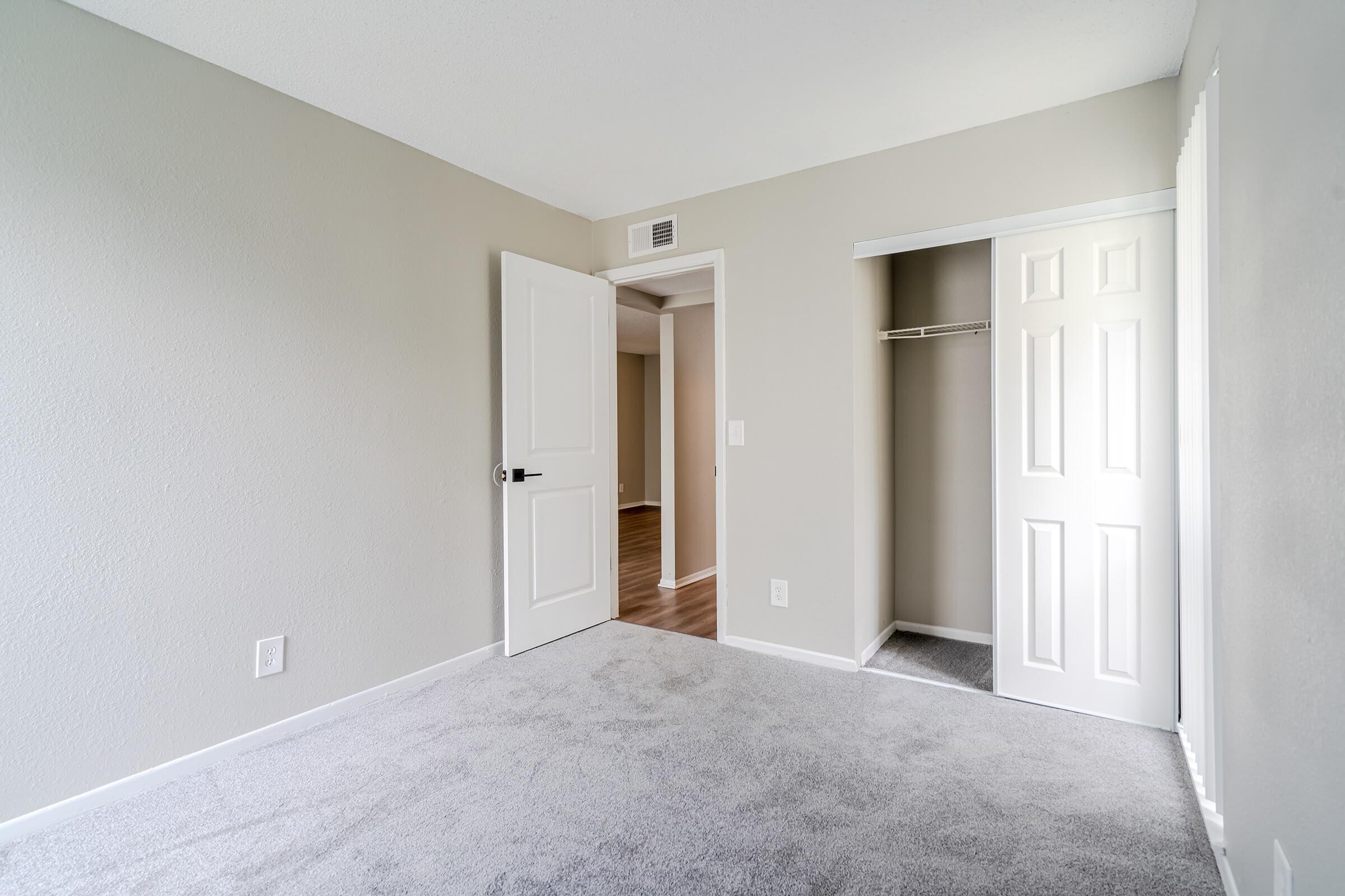 A spacious, empty room with light gray walls and carpet. One door leads to an adjacent area, while another door opens to a closet. Soft, natural light illuminates the space, creating a calm and inviting atmosphere. The floor is tidy, with no furniture present.