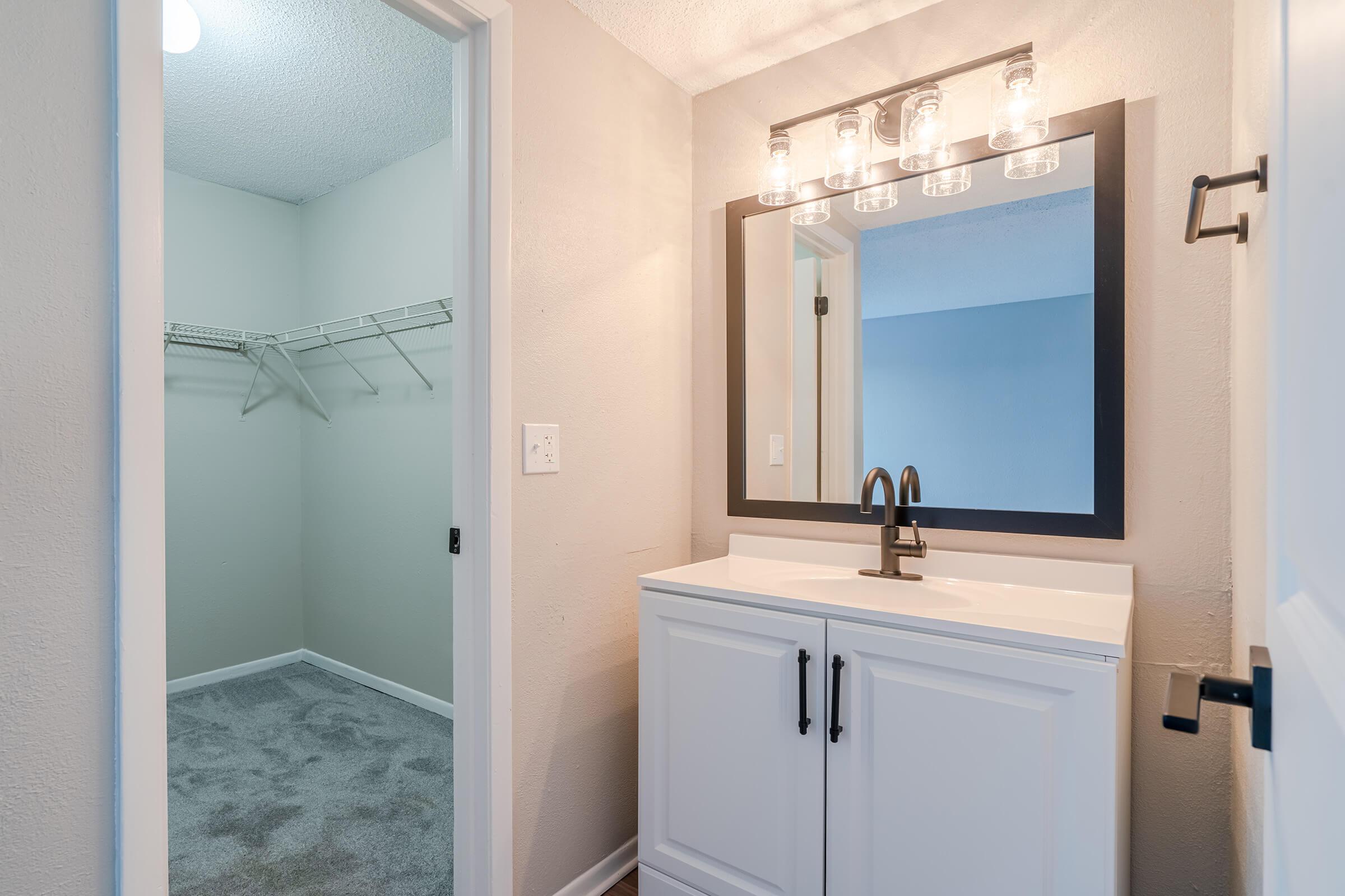 A modern bathroom featuring a white vanity with a sink, a large mirror framed in black, and stylish light fixtures above. To the left, there is a closet with a wire shelf. The walls are painted a light neutral color, and the flooring is a warm wood tone. The space appears bright and well-maintained.