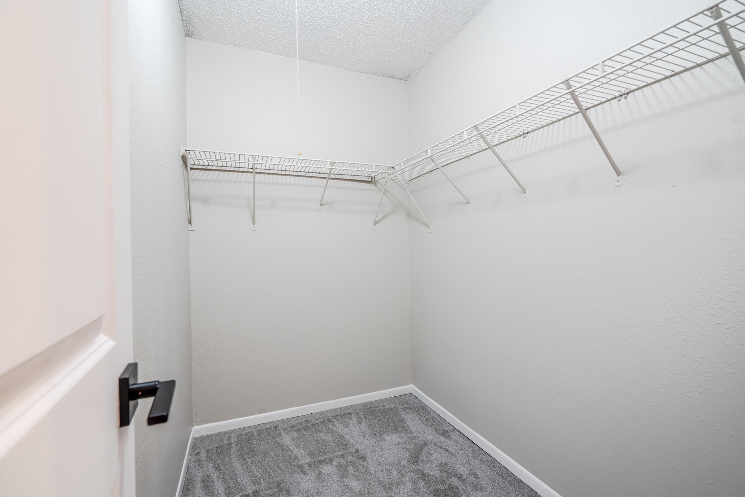 A small, empty closet with light gray walls and carpet. It features a metal wire shelf mounted on one wall, providing space for storage. The door has a modern black handle, and the area is well-lit with a ceiling light fixture.