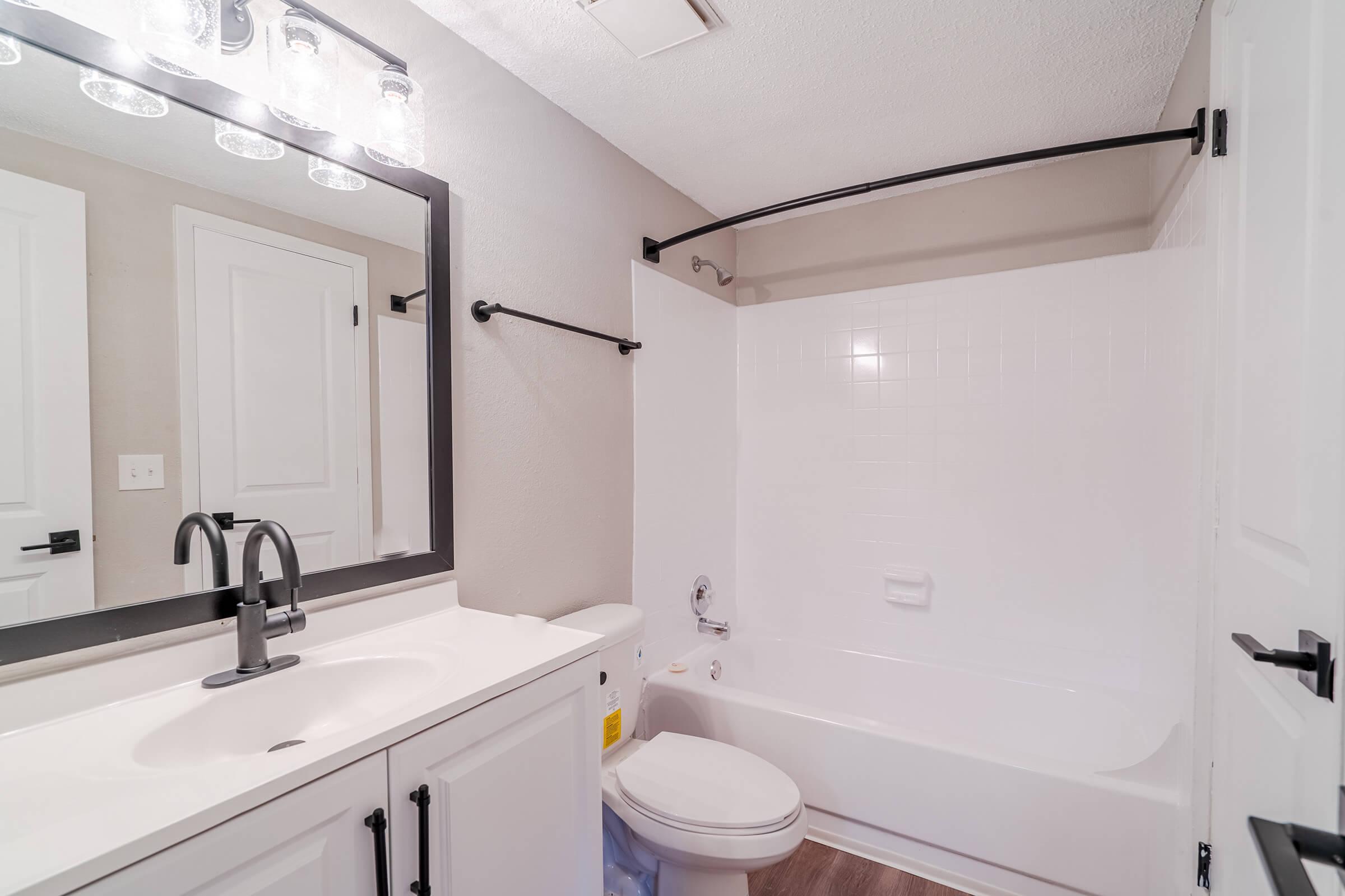 A modern bathroom featuring a white bathtub with a shower, a black metal shower rod, and a stylish black mirror above a white sink cabinet. The walls are painted in a neutral tone, and there is a towel rack and a toilet visible, along with a door that opens into the space.