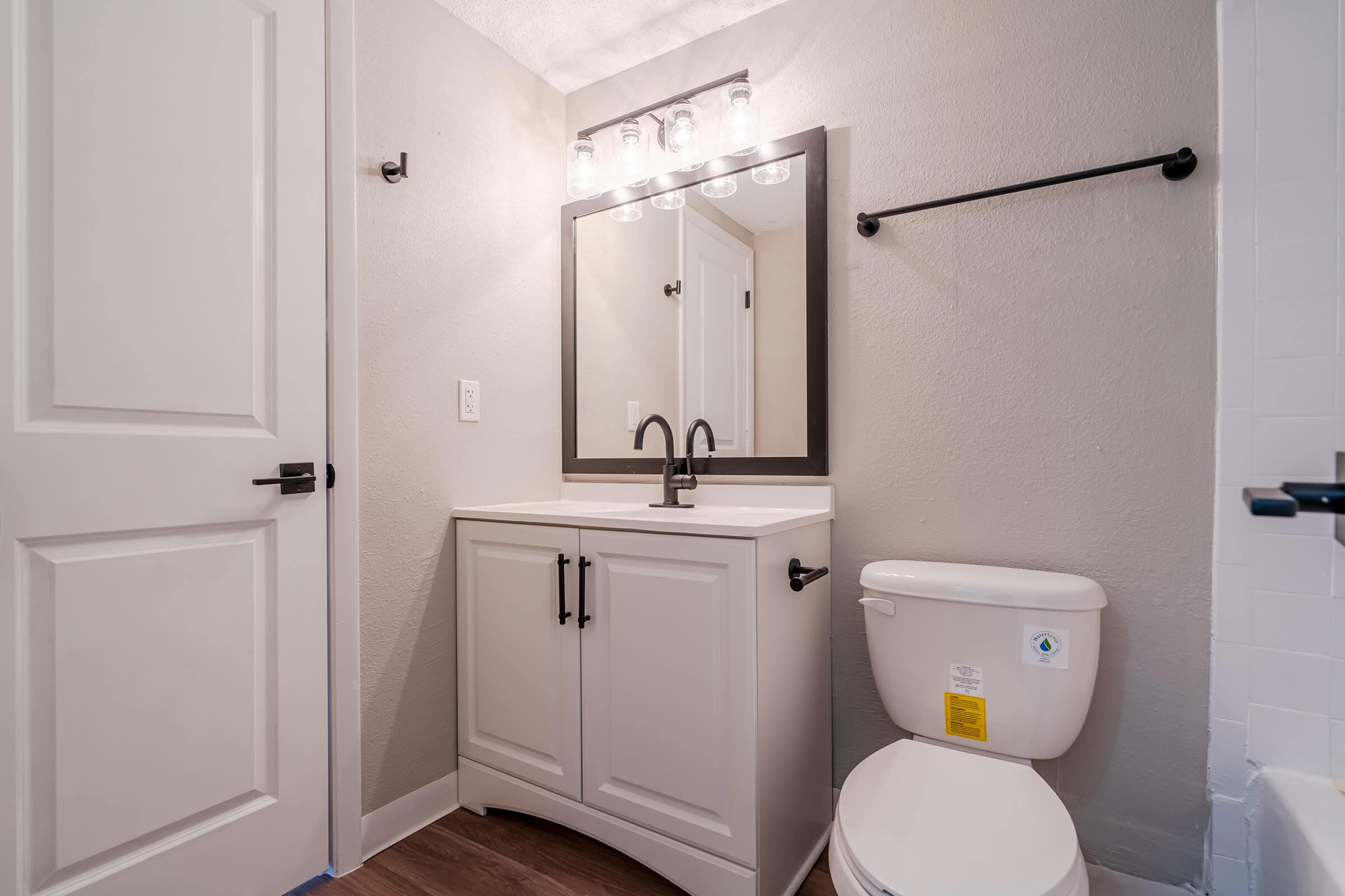 A modern bathroom featuring a white vanity with black handles beneath a large rectangular mirror. The space is illuminated by four pendant lights. There is a toilet beside the vanity, and the walls are painted in a light gray color. The flooring is wood-like, adding warmth to the design.