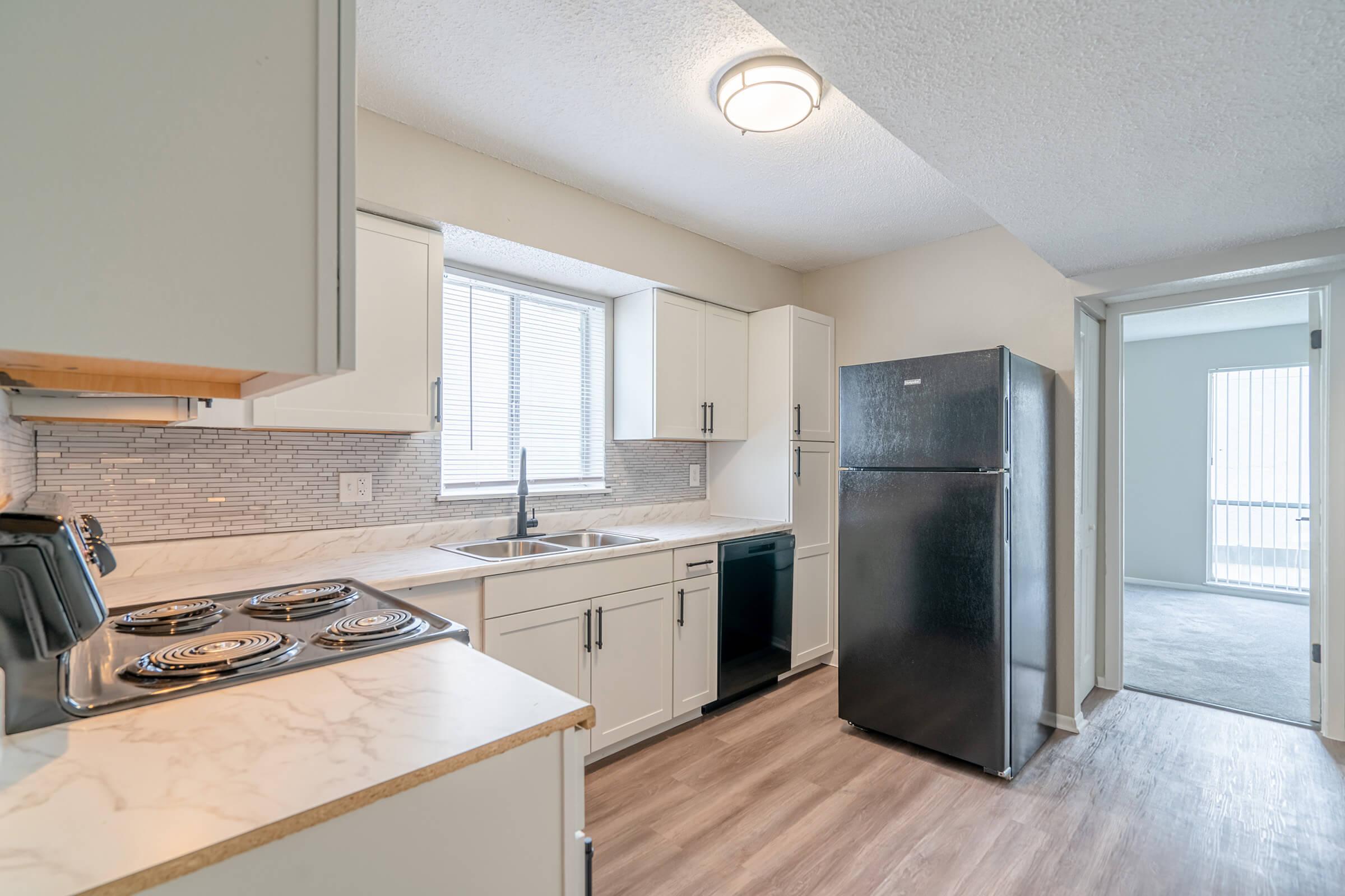 Modern kitchen featuring white cabinetry, a stainless steel refrigerator, an electric stove with four burners, and a sink. Natural light enters through a window above the sink, and the flooring is a light wood laminate. A doorway leads to an adjacent room.