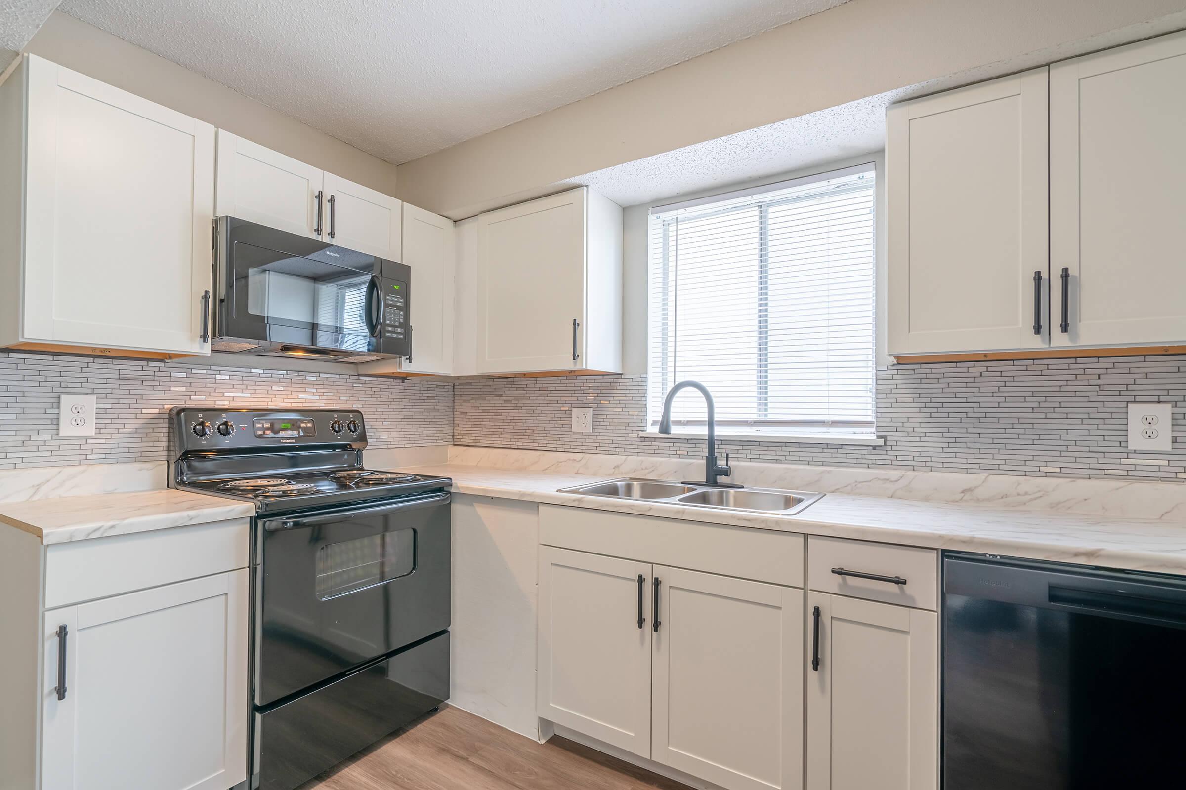 A modern kitchen featuring white cabinets, a black oven and microwave, stainless steel sink, and a window above the sink. The countertops are light-colored, and the backsplash consists of gray and white tile. The floor is a wood-like laminate, creating a sleek and inviting cooking space.