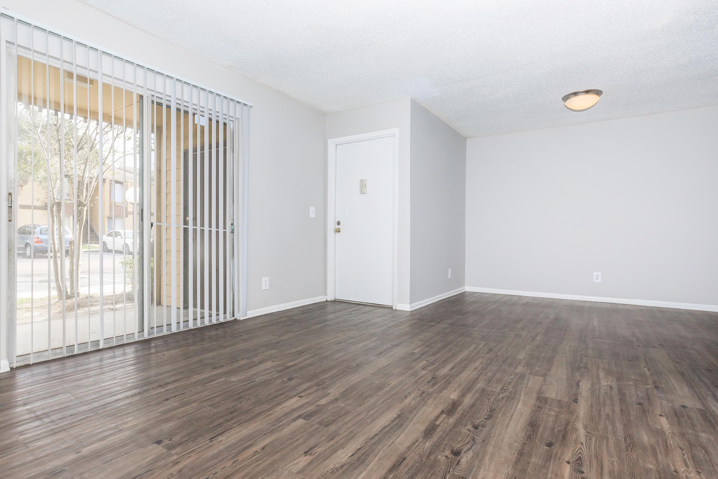 A spacious, empty room with light-colored walls and a textured ceiling. There's a door on the right leading outside and a large window with vertical blinds on the left. The floor is a dark wood laminate, creating a modern and clean look in the interior space.