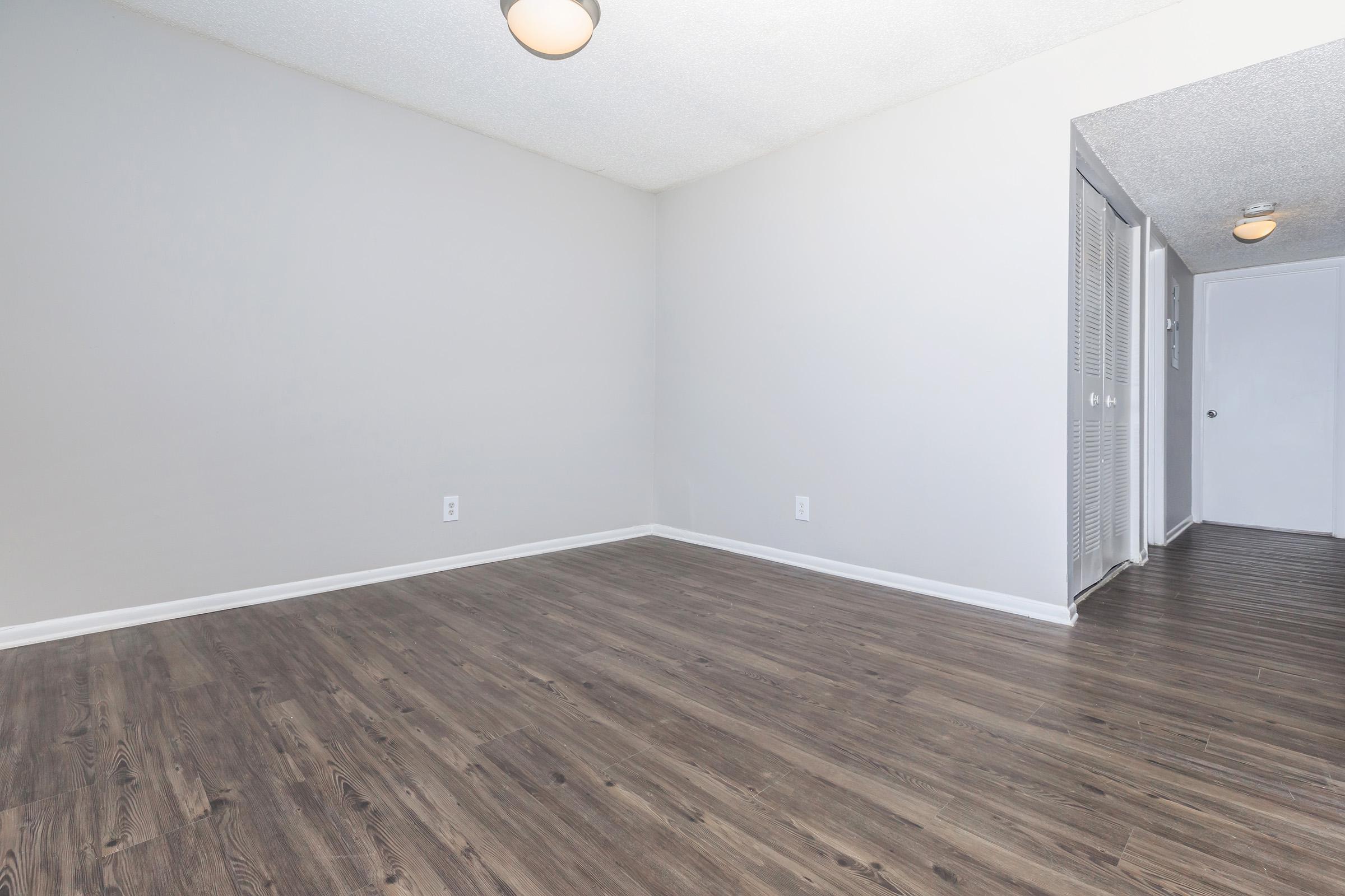 A vacant room with light gray walls and dark laminate flooring. The ceiling features a round light fixture. On the right, there are closed doors leading to a hallway, while the room appears spacious and unfurnished, creating a minimalist environment.