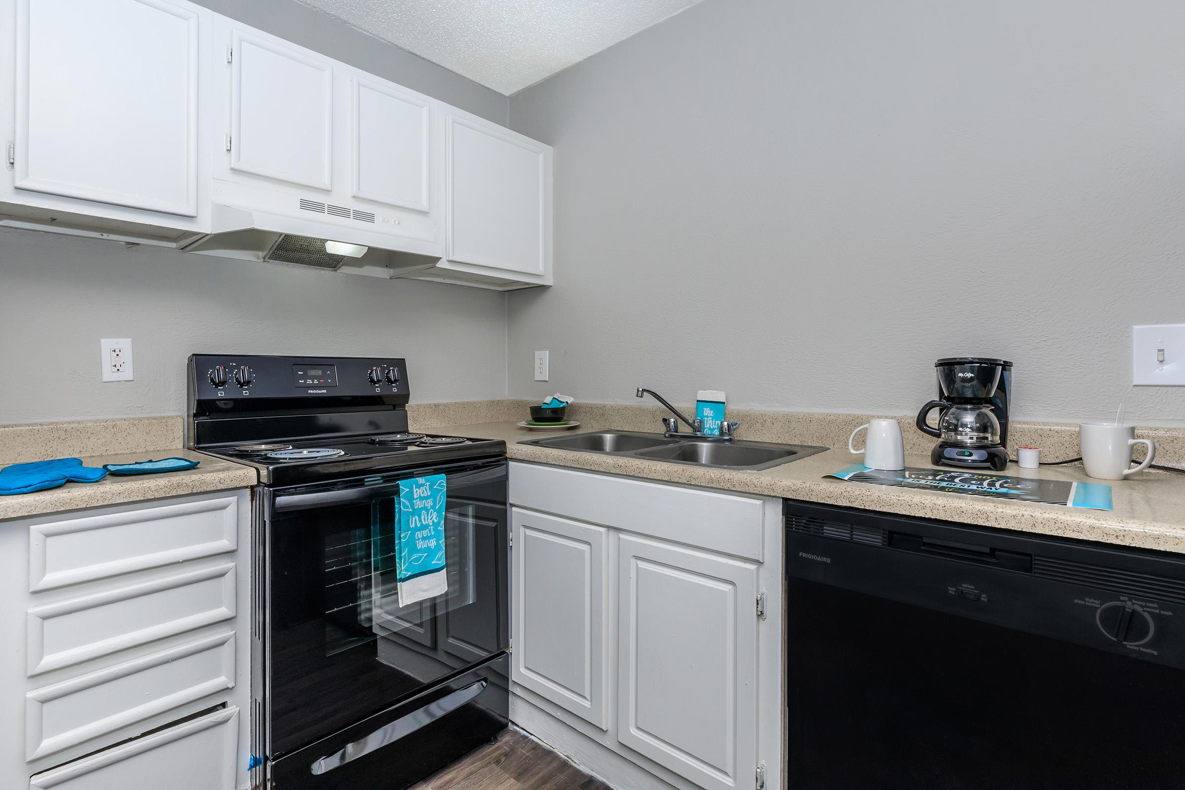 A modern kitchen featuring white cabinetry, a black stove and oven, a sink, and a coffee maker. The countertops are light-colored, and a few blue kitchen towels and utensils are visible. The walls are painted gray, creating a clean and contemporary look.