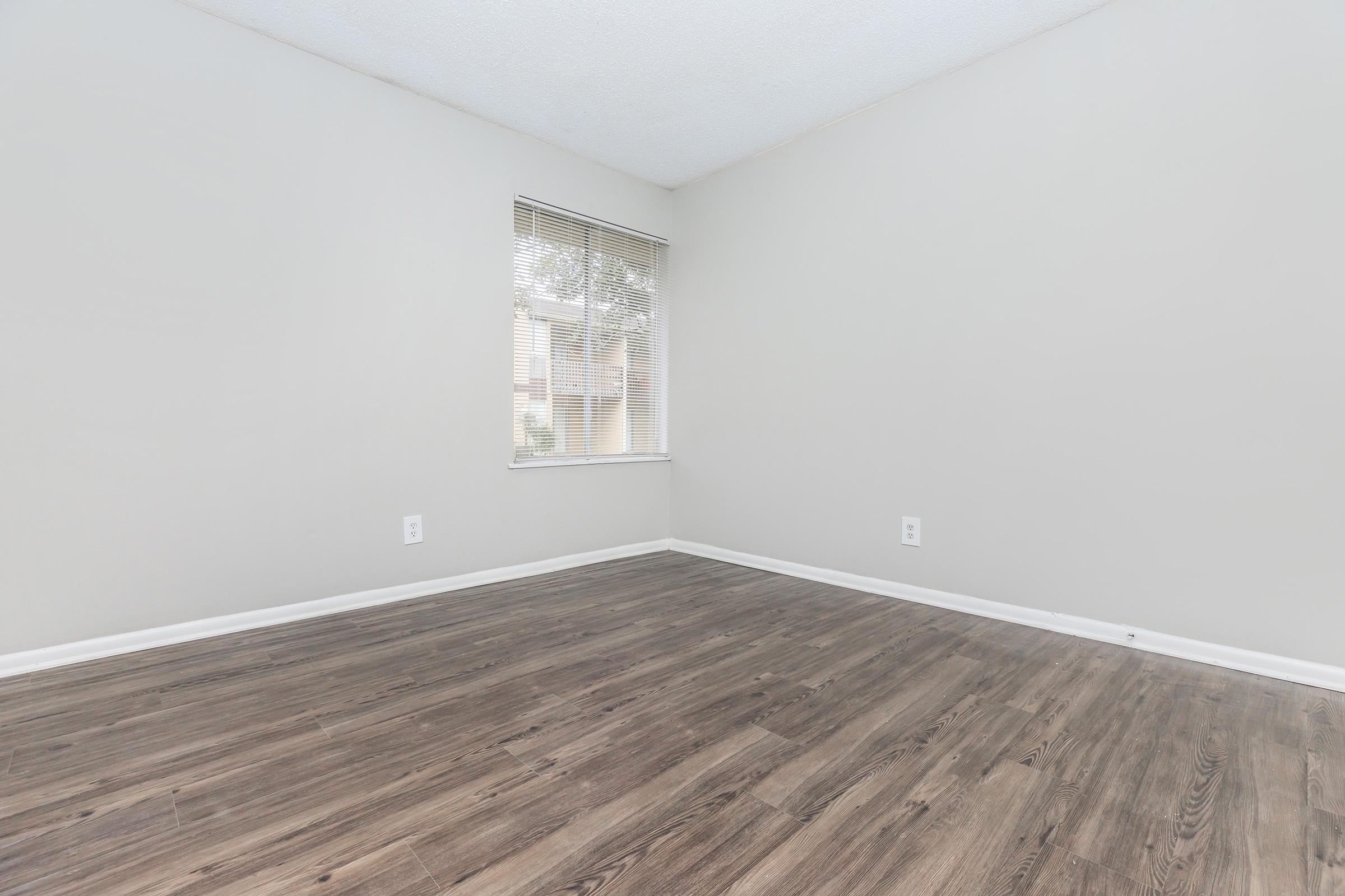 A light-colored room with beige walls and wooden flooring. There is a window with blinds on one wall, allowing natural light to enter. The room is empty and spacious, featuring minimal decor, creating a calm and neutral atmosphere.