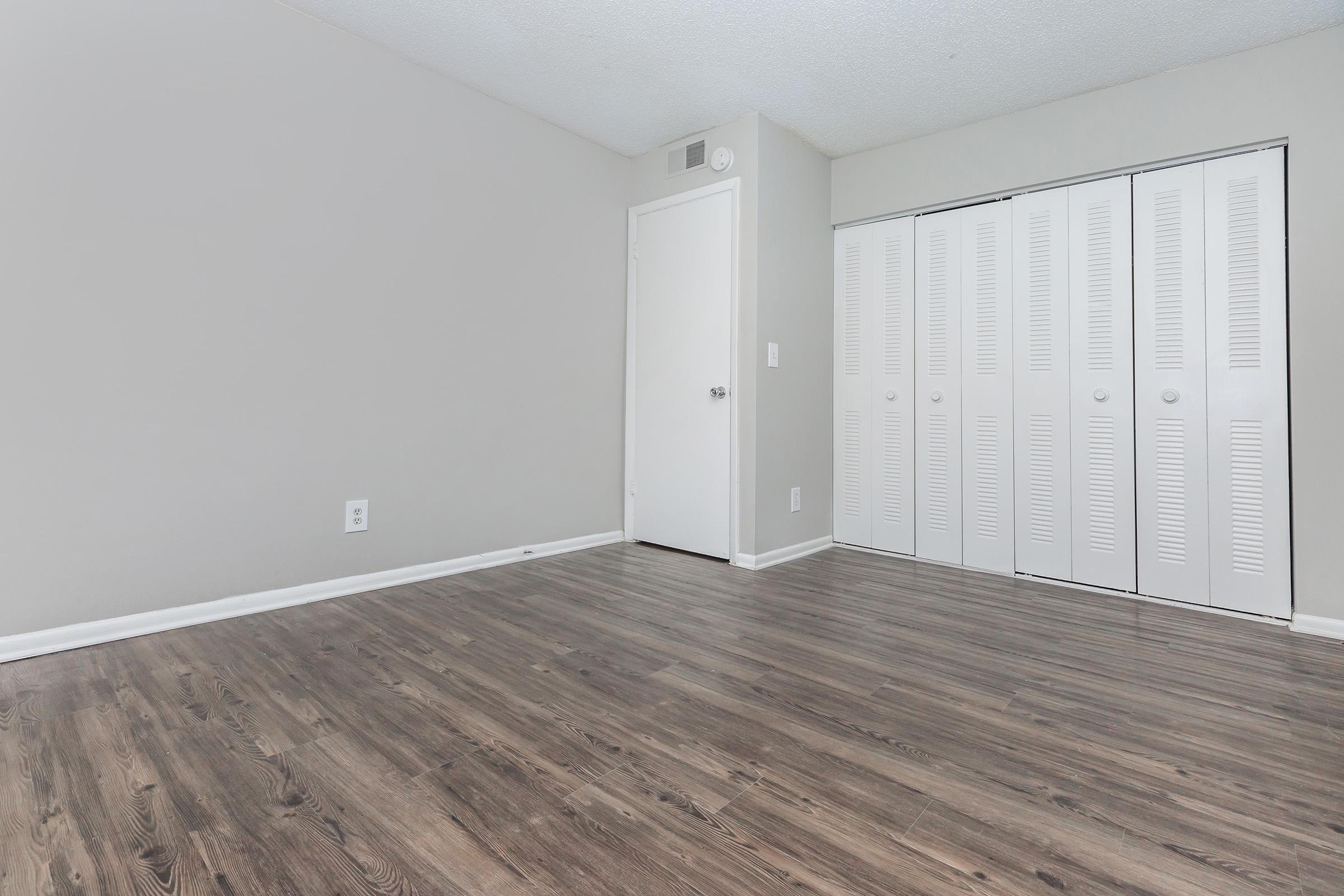 A clean, empty room featuring light-colored walls and a modern laminate floor. Empty closet with white louvered doors on the right side. A closed white door is visible on the left. The space is well-lit, offering a minimalist and spacious feel.