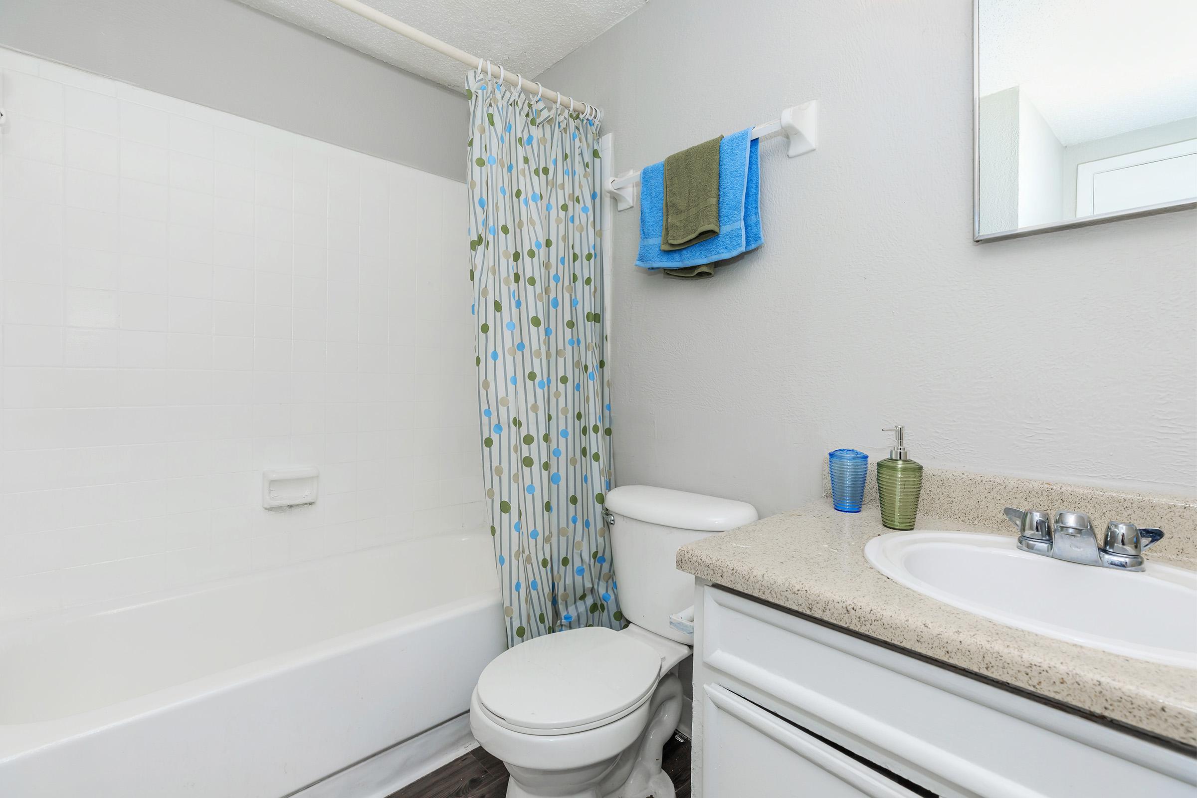 A clean and simple bathroom featuring a bathtub with a shower curtain having a green and blue polka dot design. There's a white toilet, beige countertop with a sink, and a large mirror. Two towels hang on a rack above the toilet, and small decorative items are on the counter.