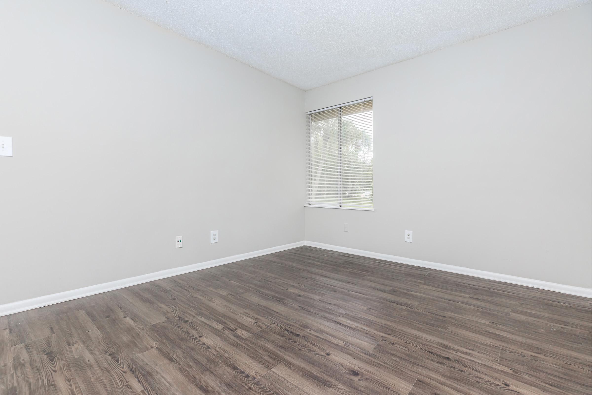 Empty room with light gray walls and brown laminate flooring. A single window with blinds is visible, allowing natural light to enter. The space is unfurnished, creating a clean and minimalistic appearance.