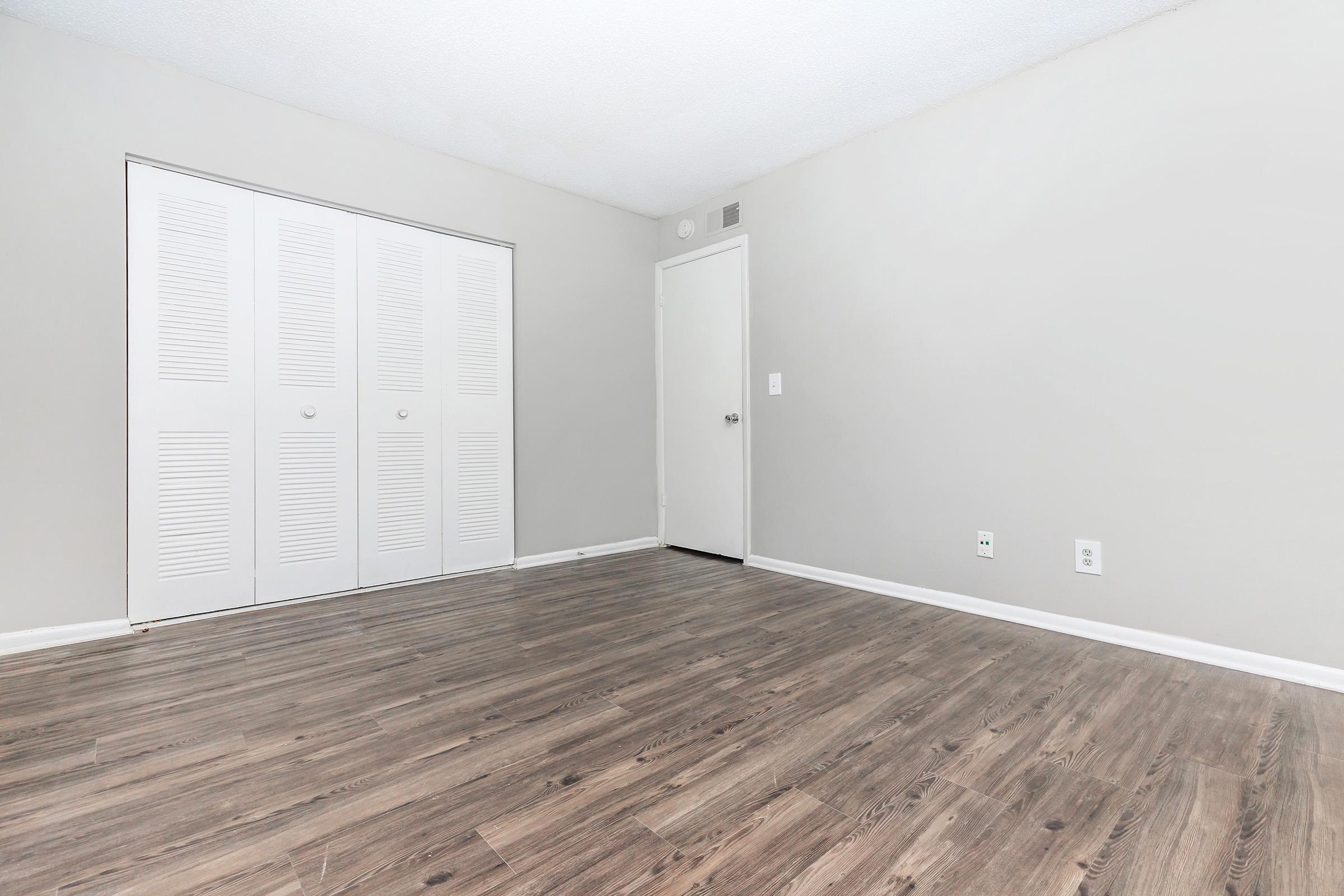 A spacious, empty room with light gray walls and a wooden floor. Two white closet doors with horizontal slats are on one wall, while a plain white door is on another wall. The room is well-lit and has an open, minimalist feel with no furniture present.