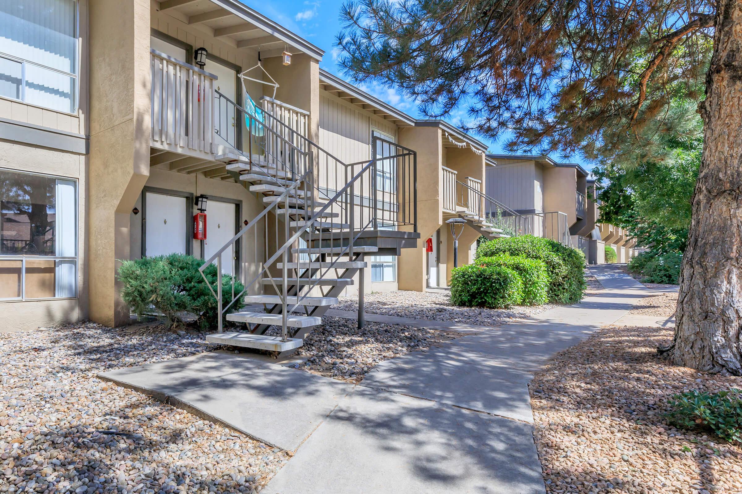 A view of an apartment complex featuring two-story buildings with stairs leading to upper balconies. The pathway in the foreground is lined with pebble landscaping and greenery, providing a well-maintained outdoor environment. Bright blue sky above adds to the pleasant atmosphere.