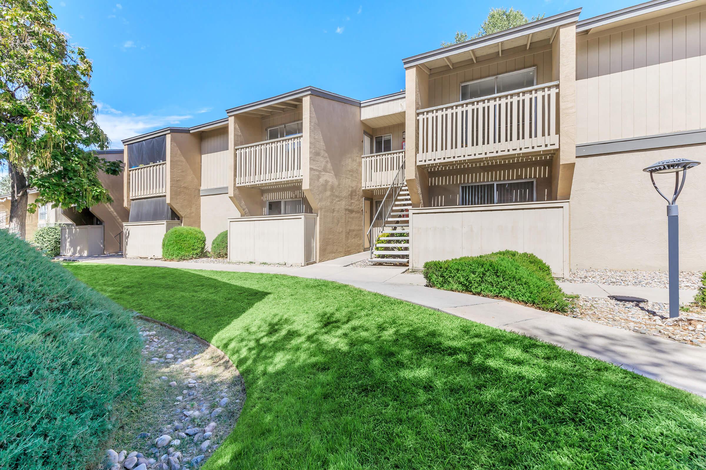 Two-story apartment building with beige exterior and wooden balconies. Lush green grass and shrubs surround a curved pathway leading to the entrance. The scene is bright with clear blue skies, creating a welcoming atmosphere. A lamp post is visible along the path.