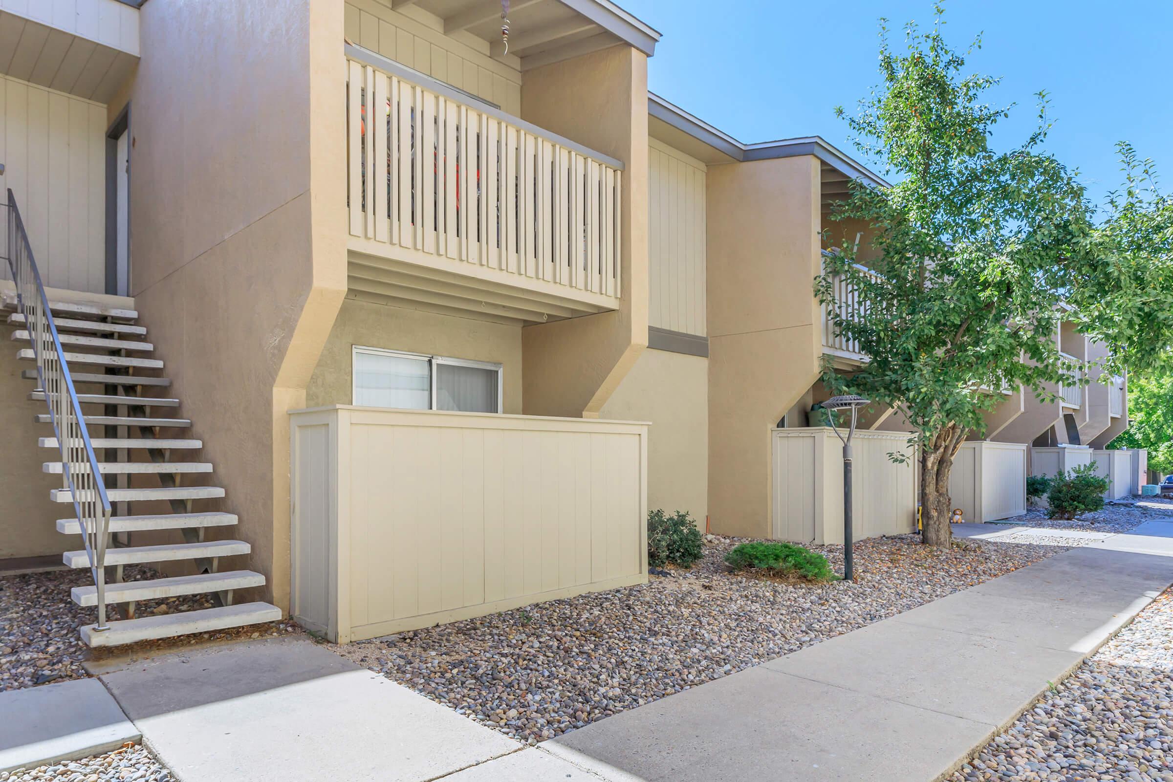 A view of a multi-unit residential building featuring beige exterior walls and balconies. A set of stairs leads to the upper-level units on the left, while the landscaped area includes gravel, shrubs, and a pathway. The scene is bright and welcoming under a clear blue sky.