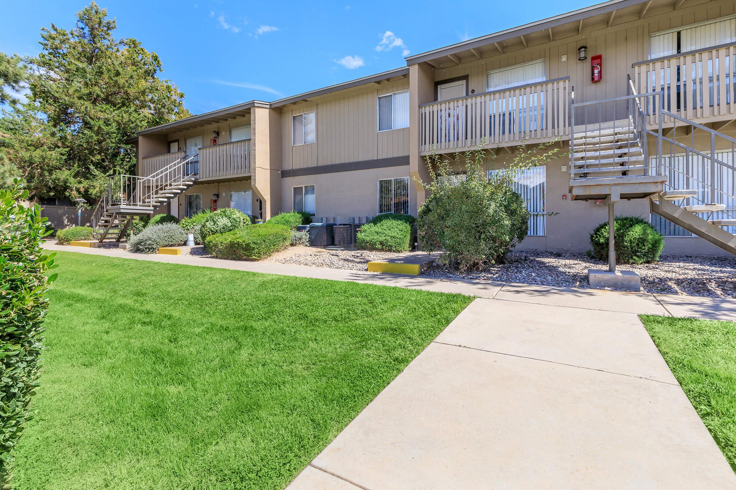 A view of an apartment complex featuring two-story buildings with staircases, surrounded by green lawns and shrubs. The pathway leads through the landscaped area, with clear blue skies in the background.