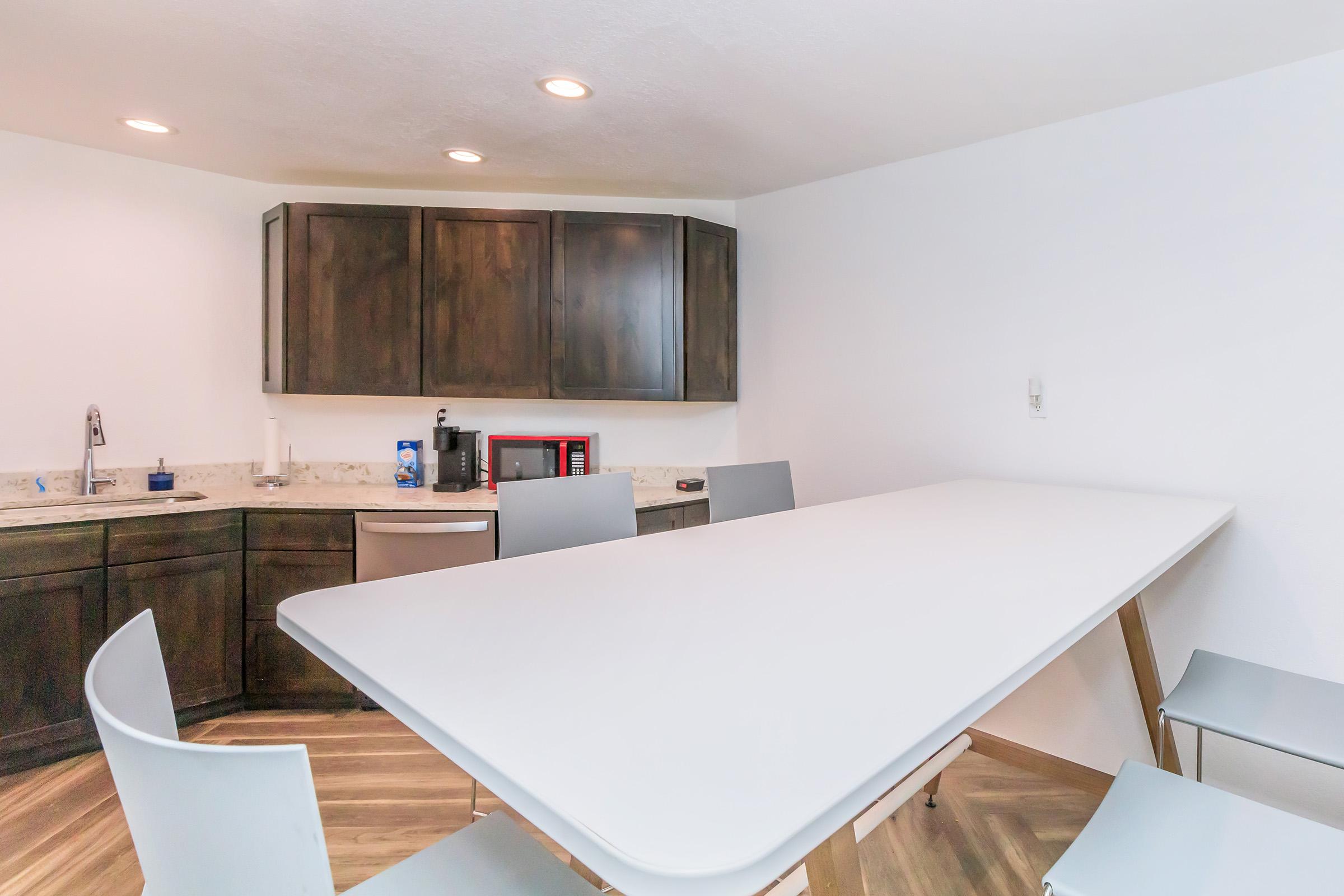 A modern kitchen with a sleek, white table surrounded by four gray chairs. The kitchen features dark wooden cabinets, a stainless steel dishwasher, and a countertop with a coffee maker and microwave. The walls are painted white, creating a bright and open atmosphere.