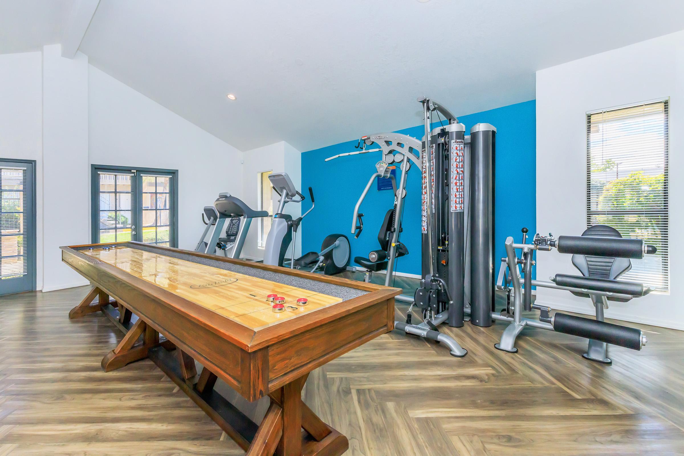 A modern home gym featuring a variety of exercise equipment, including a treadmill, weight machine, and exercise bikes. There's also a wooden shuffleboard table in the foreground, adding a recreational element to the space, with large windows allowing natural light to brighten the room.