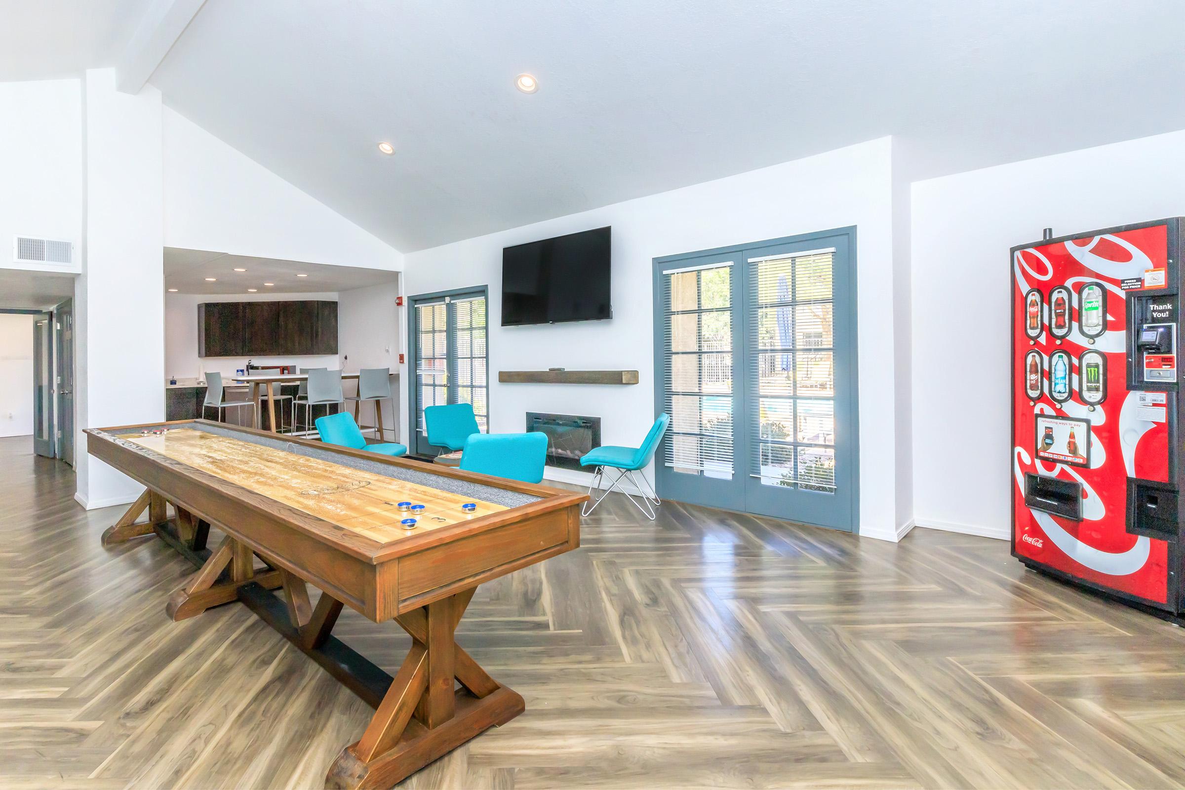 A spacious indoor lounge area featuring a large wooden table, bright blue chairs, a flat-screen TV mounted on the wall, and a vending machine. Large windows allow natural light to fill the space, with a cozy fireplace visible in the background. The decor is modern and inviting.
