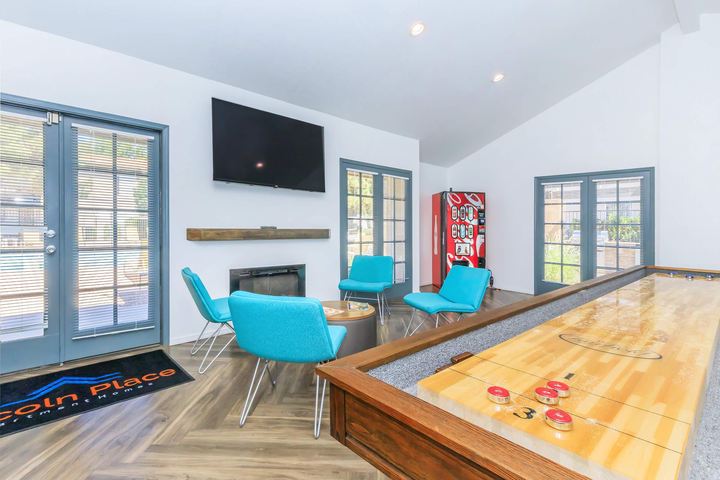 A modern lounge area featuring a large flat-screen TV mounted on the wall, a wooden table with a shuffleboard, and turquoise chairs around a small table. Large windows allow natural light to illuminate the space. A vending machine is visible in the corner. The flooring consists of wood laminate.