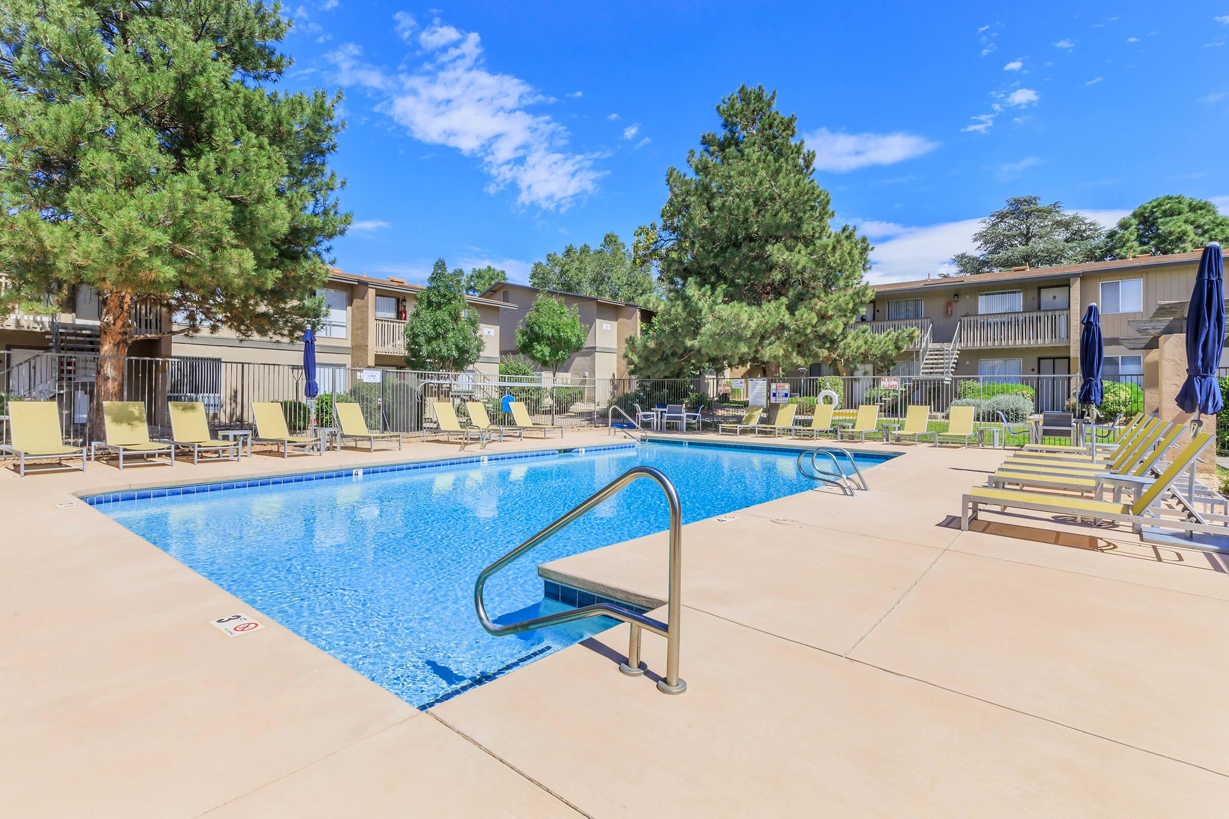 A clear blue swimming pool surrounded by lounge chairs under sunny skies. Green trees provide shade, and there are two buildings in the background, creating a relaxing outdoor space.