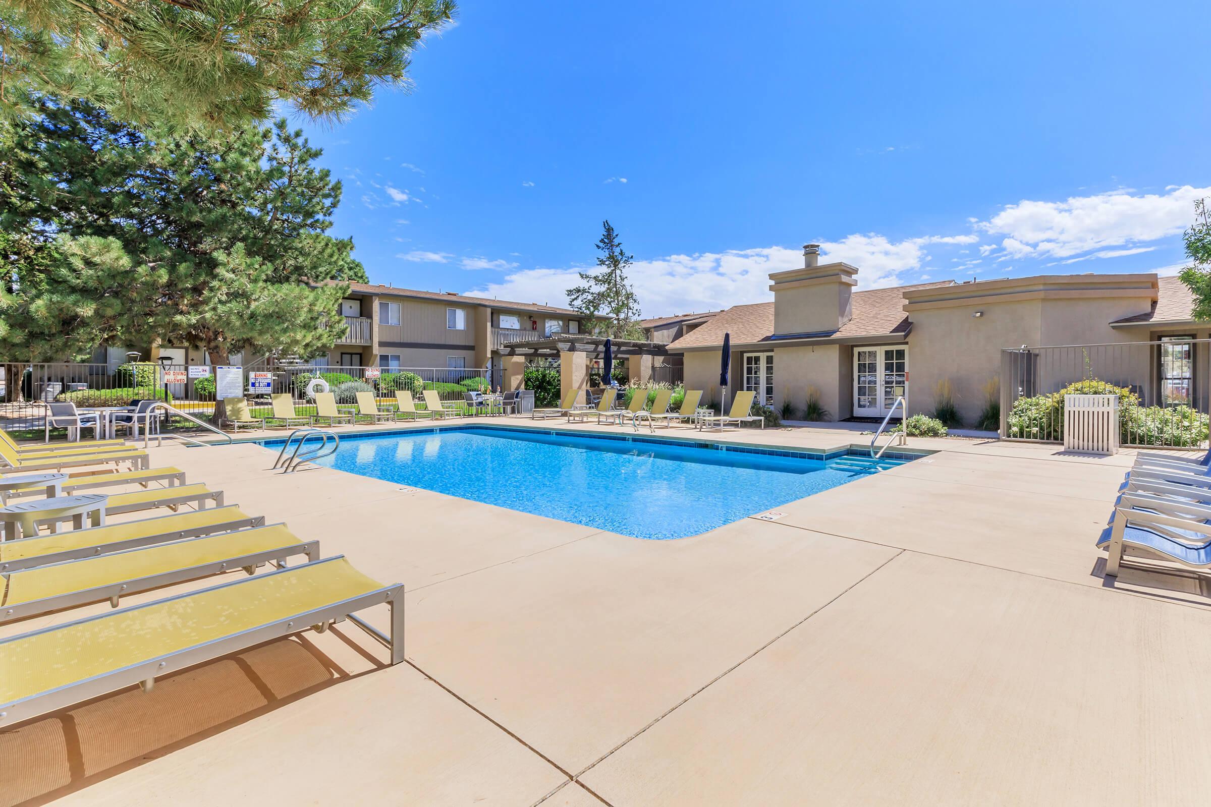 A bright outdoor pool area featuring a clear blue pool surrounded by lounge chairs. Lush green trees line the space, and there are residential buildings in the background under a clear blue sky. The setting is inviting and perfect for relaxation.