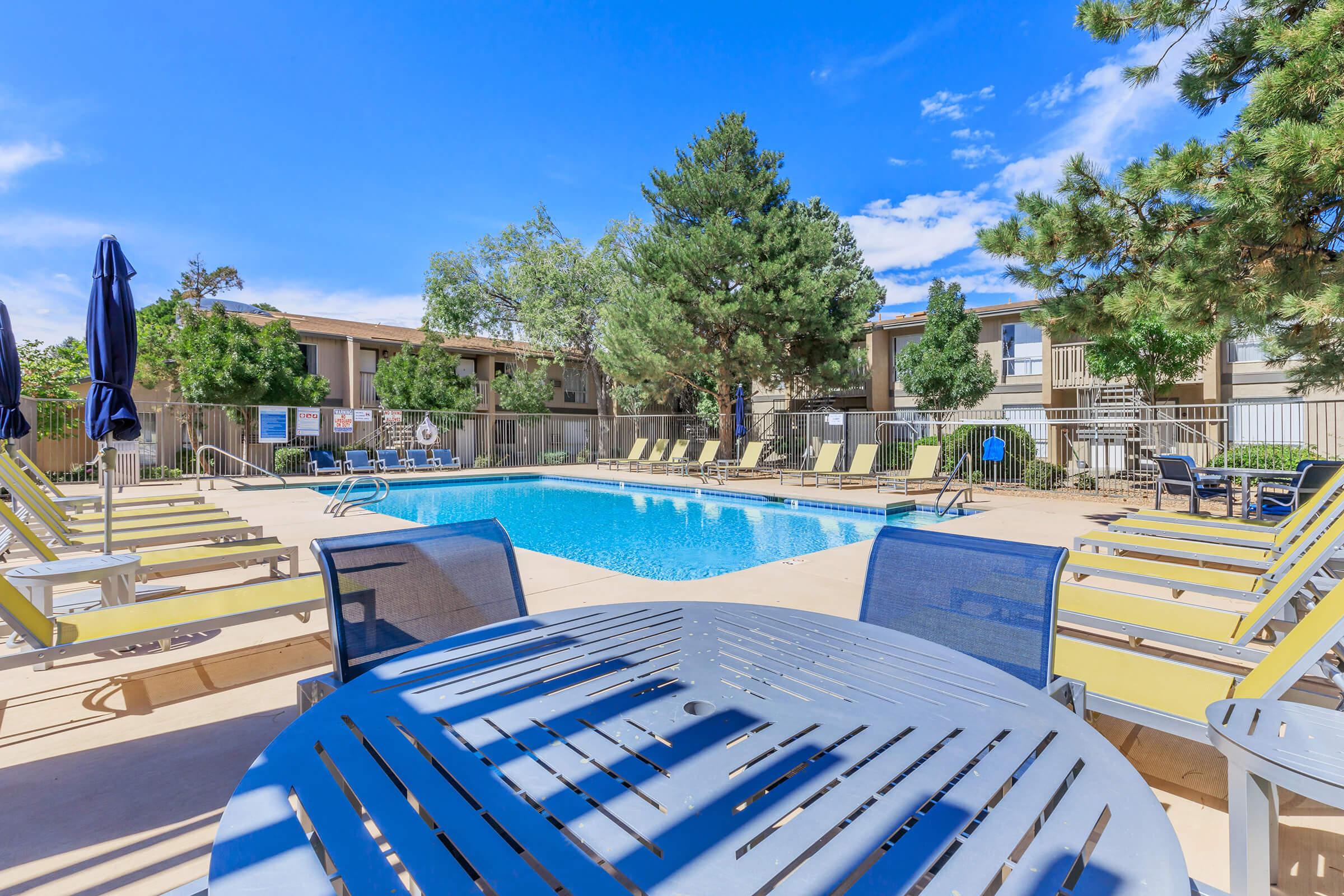 A bright, sunny pool area featuring a rectangular swimming pool surrounded by lounge chairs and umbrellas. In the background, there are trees and a multi-story building, with a clear blue sky overhead. A table with chairs is in the foreground, inviting for relaxation and socializing.