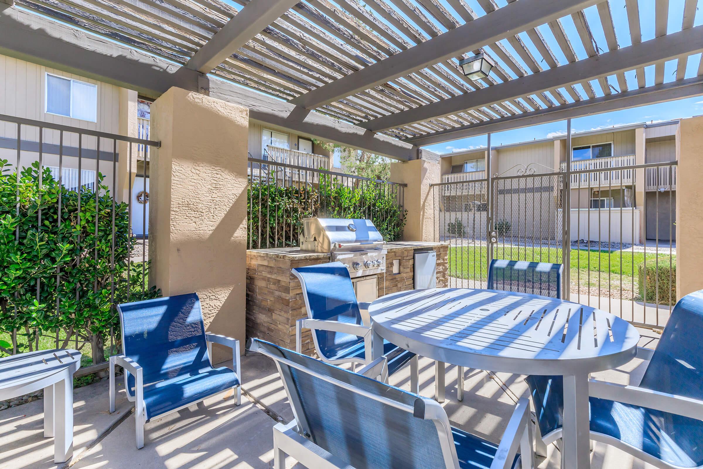 Outdoor patio area featuring a stone barbecue grill under a shaded pergola. There are several blue chairs and a round table arranged around the space. The area is enclosed by a metal gate, with green foliage visible in the background, creating a welcoming and relaxing atmosphere.