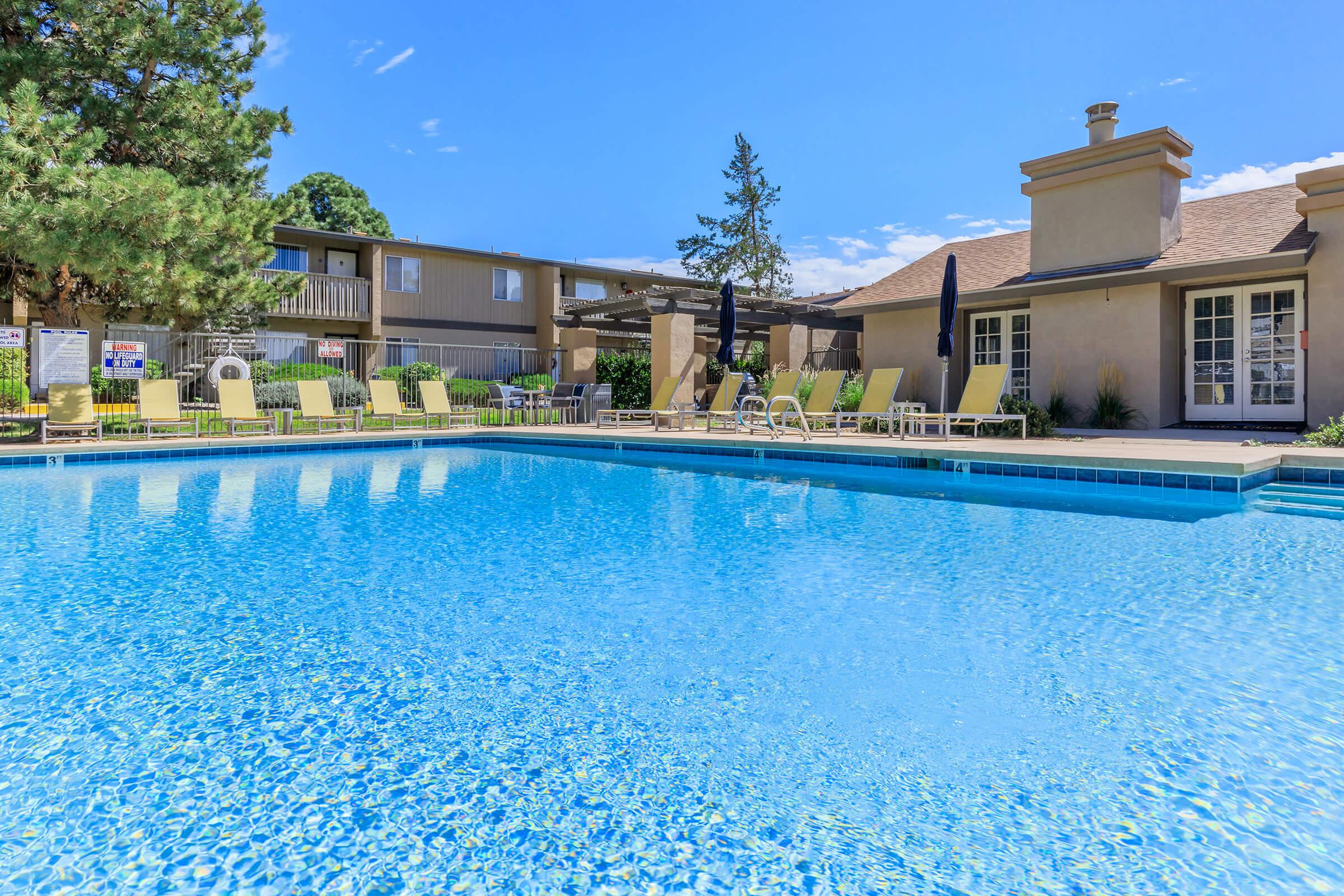 A bright and inviting outdoor swimming pool surrounded by lounge chairs and shaded areas. Lush greenery and a clear blue sky provide a serene backdrop, while a nearby building offers additional amenities. The scene is perfect for relaxation and leisure.