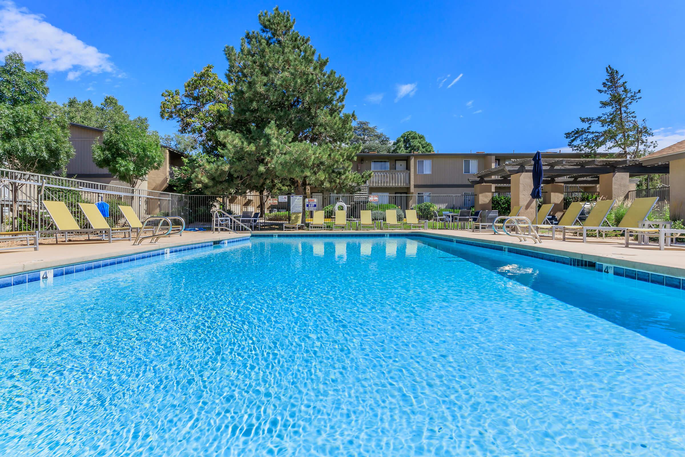 A clear swimming pool surrounded by lounge chairs under a bright blue sky. Lush green trees and well-maintained landscaping are visible in the background, creating a relaxing outdoor atmosphere. The pool's surface reflects the sunlight, adding to the inviting scene.