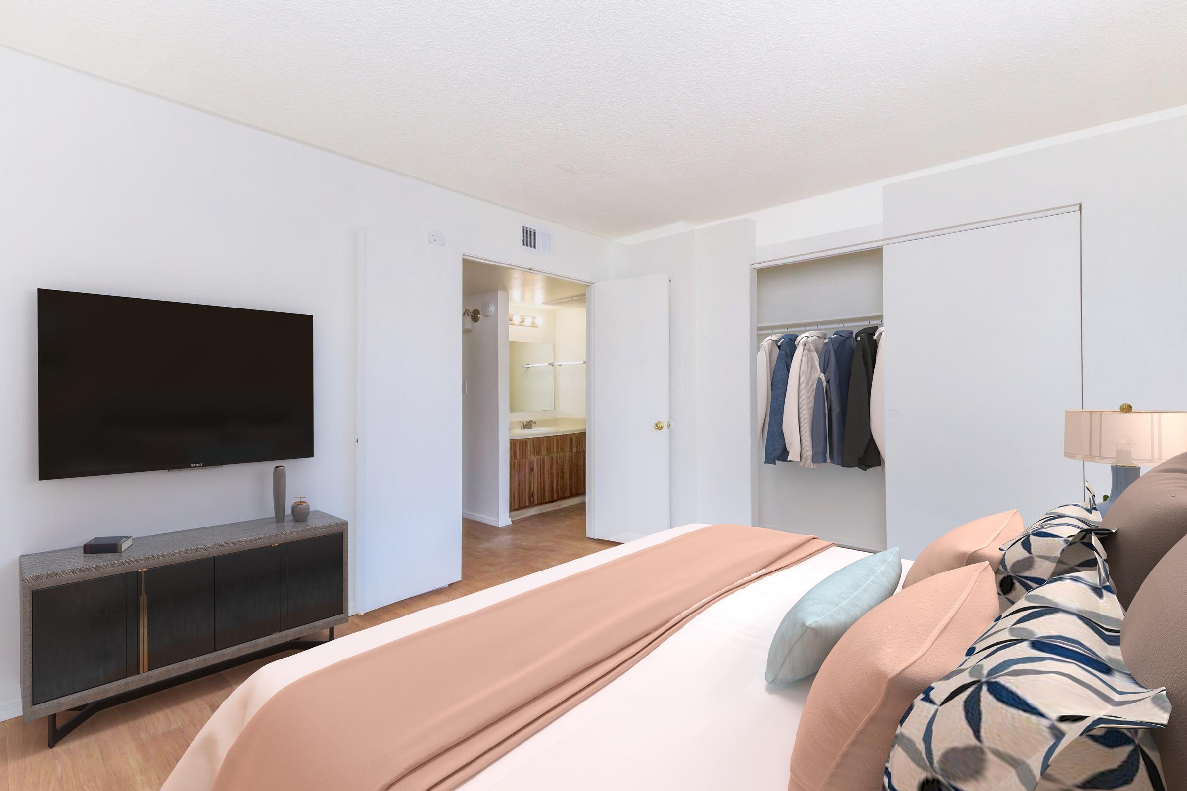 A modern bedroom featuring a neatly made bed with decorative pillows, a bedside lamp, and a contemporary television on a dresser. A closet is visible in the background with hangers holding clothing, and a doorway leads to a bathroom area. The walls are painted in a light color, enhancing the spacious feel.