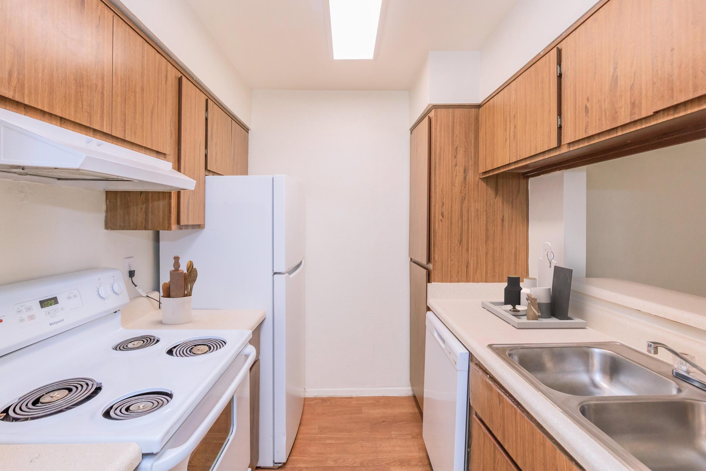 A modern kitchen featuring light wood cabinetry, a white refrigerator, and a white stove with four burners. There's a double sink with a soap dispenser, and the countertops are light-colored. The room is well-lit with natural light coming from a ceiling fixture. The floors are a warm, light wood tone.
