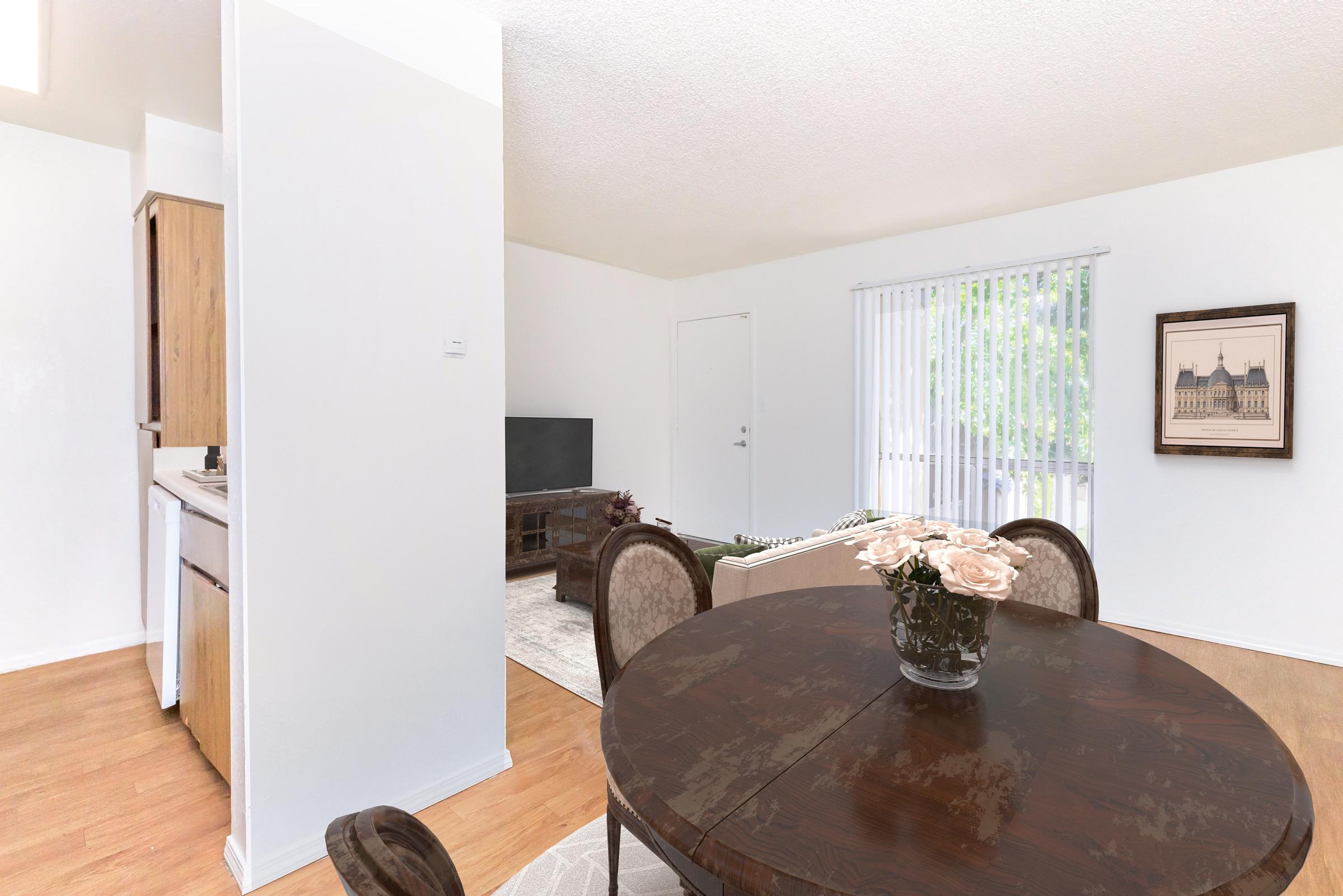 A bright, open living space featuring a circular dining table with a vase of pink roses, a kitchenette visible on the left, and a cozy living area with a TV and light-colored sofa. Large windows allow natural light to fill the room, highlighting the light wood flooring and neutral wall colors.