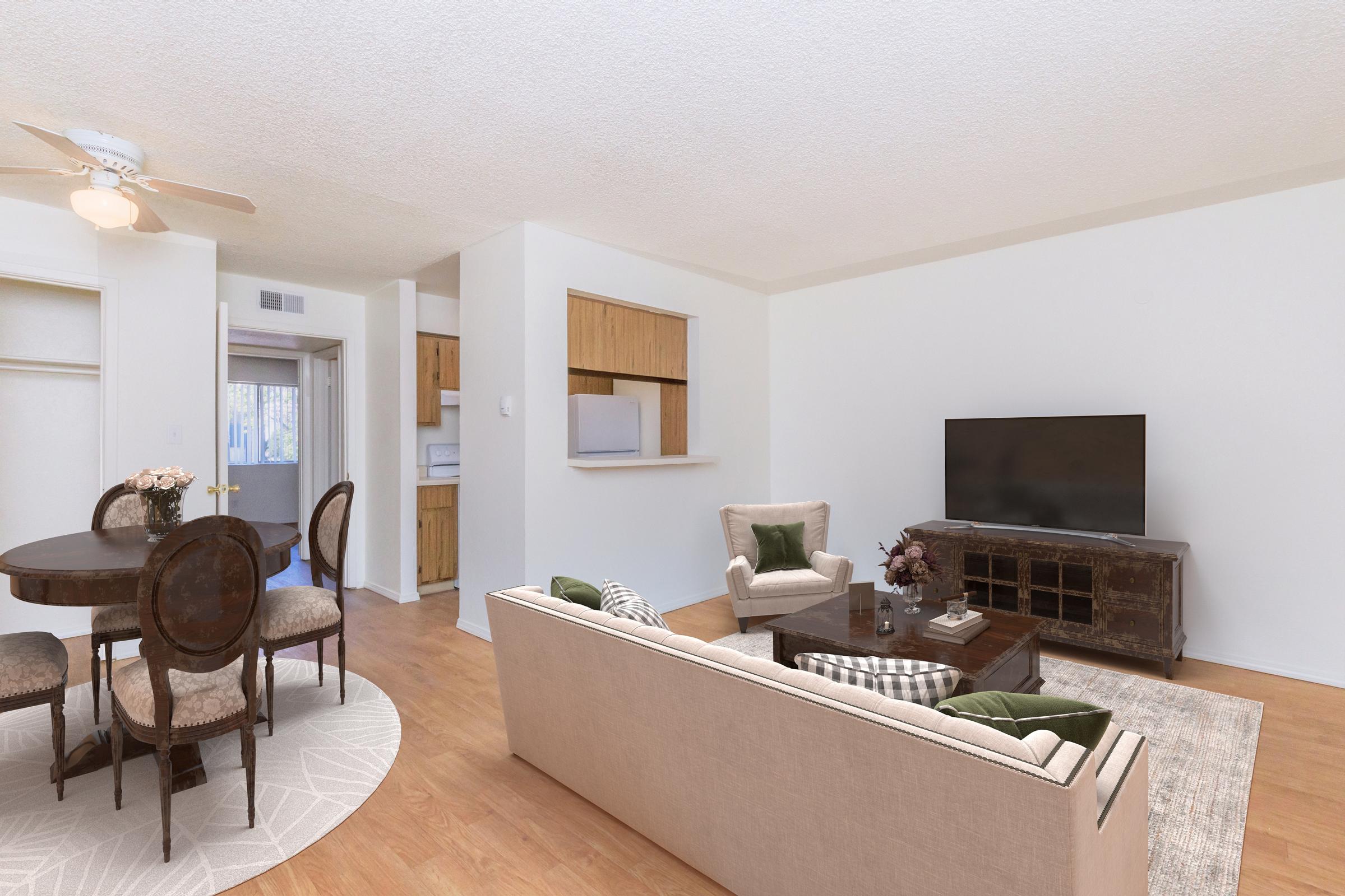 A bright, modern living room featuring a beige sofa with green cushions, a wooden coffee table, and a large TV. In the background, there is a dining area with a round table and four chairs. The space has light wood flooring and warm, neutral-colored walls, creating an inviting atmosphere.