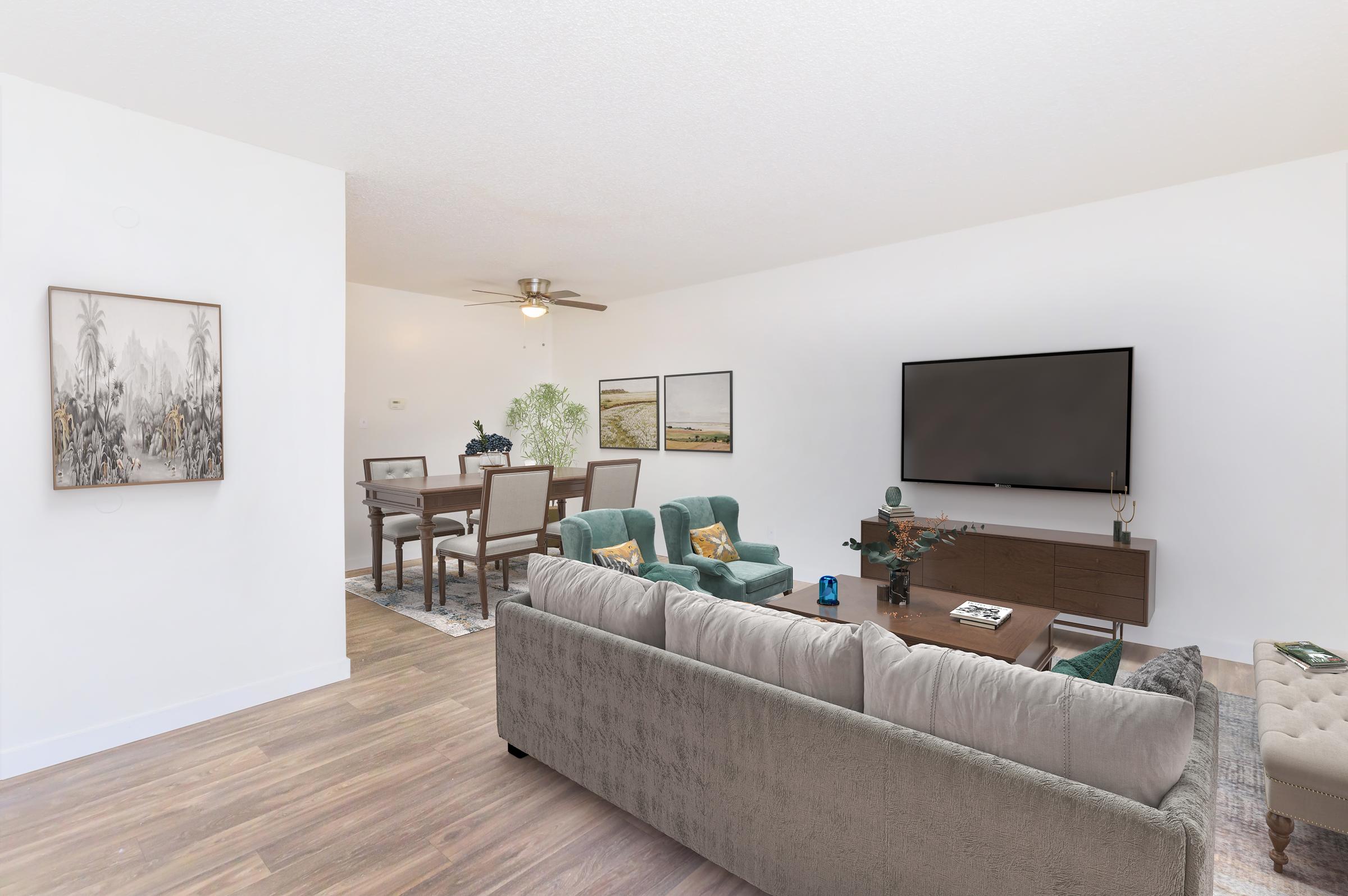 A modern living room featuring a comfortable gray sofa, green armchairs, and a wooden coffee table. A large flat-screen TV is mounted on the wall, and there are decorative plants and artwork. The space is bright and inviting, with wooden flooring and a dining area visible in the background.