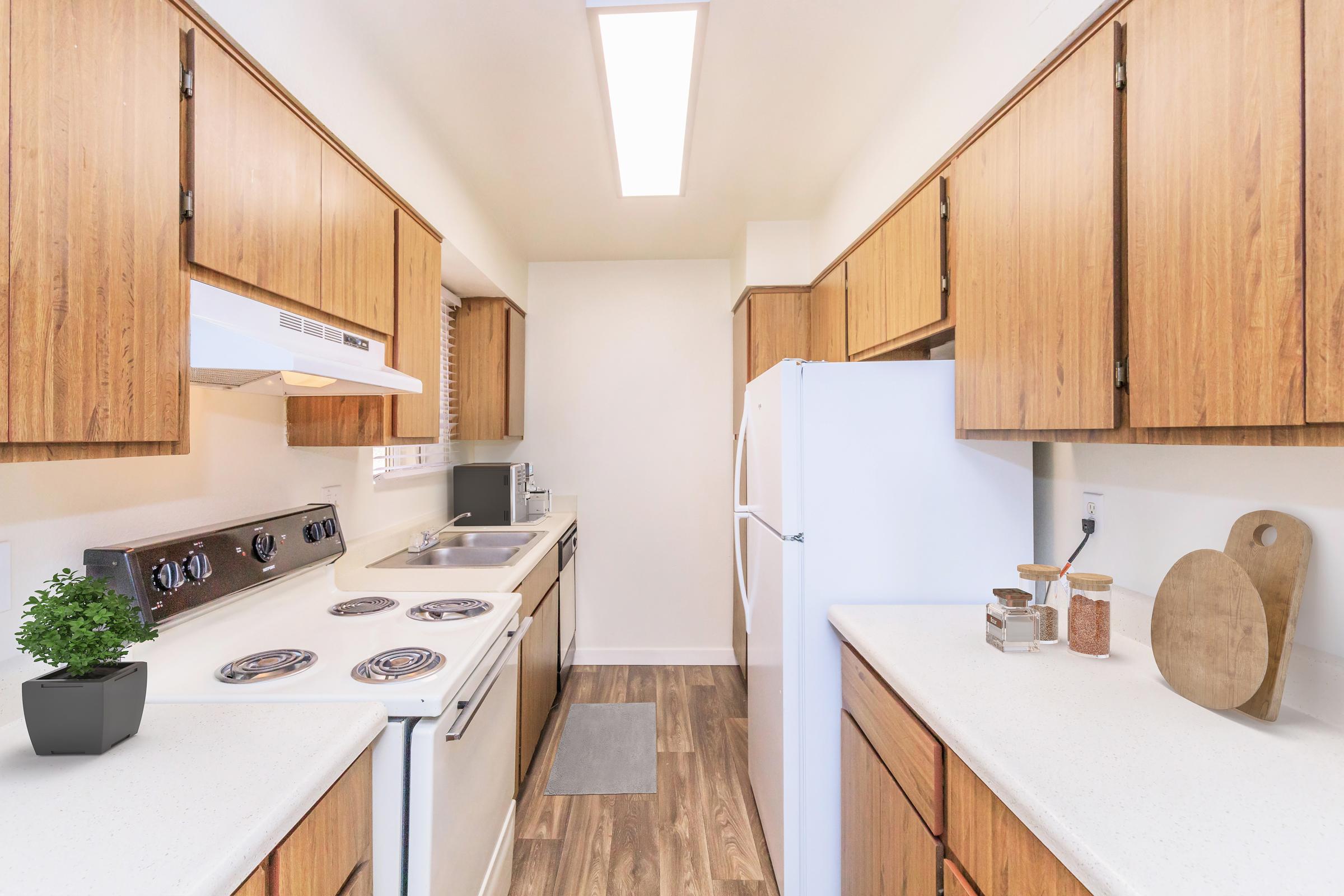 A modern kitchen with wooden cabinets, a white stove, and a refrigerator. There is a small plant on the counter, a cutting board, and jars for storage. The kitchen features a clean layout with ample counter space and natural light coming from a skylight. The floor has a wooden finish.