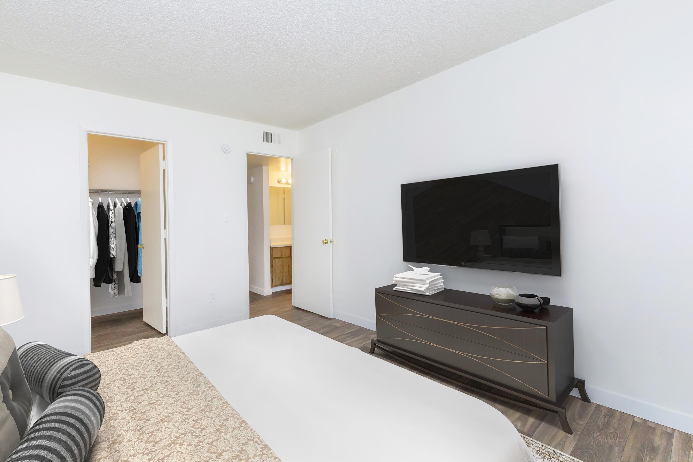 A modern bedroom featuring a neatly made bed with a folded blanket, a sleek dark wooden dresser with decorative items, and a flat-screen TV mounted on the wall. A doorway leads to a well-lit bathroom, while a closet with hanging clothes is visible to the left. The walls are painted white, creating a bright, airy feel.