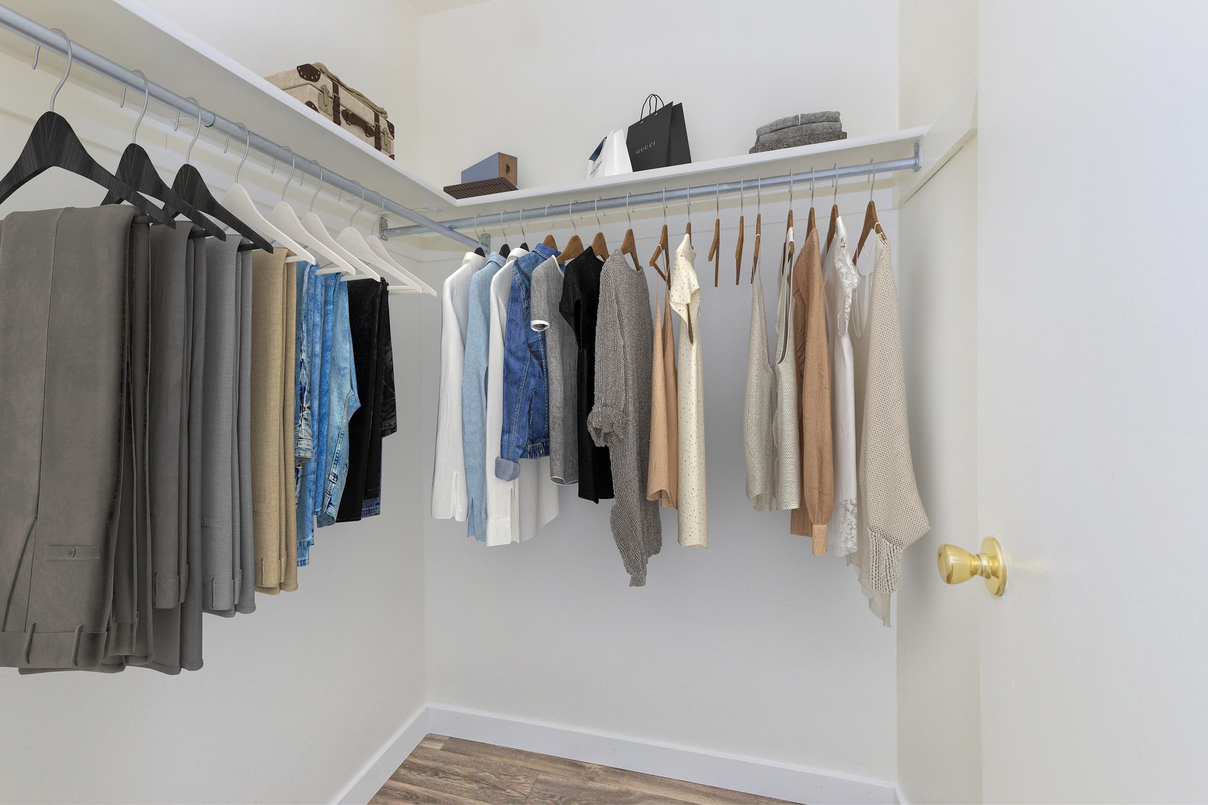 A neatly organized closet featuring various clothing items on hangers. The left side has a row of pants in shades of grey, while the right side displays shirts and tops in neutral and light colors. Accessories and folded items are visible on shelves above, with a door partially open revealing the interior.