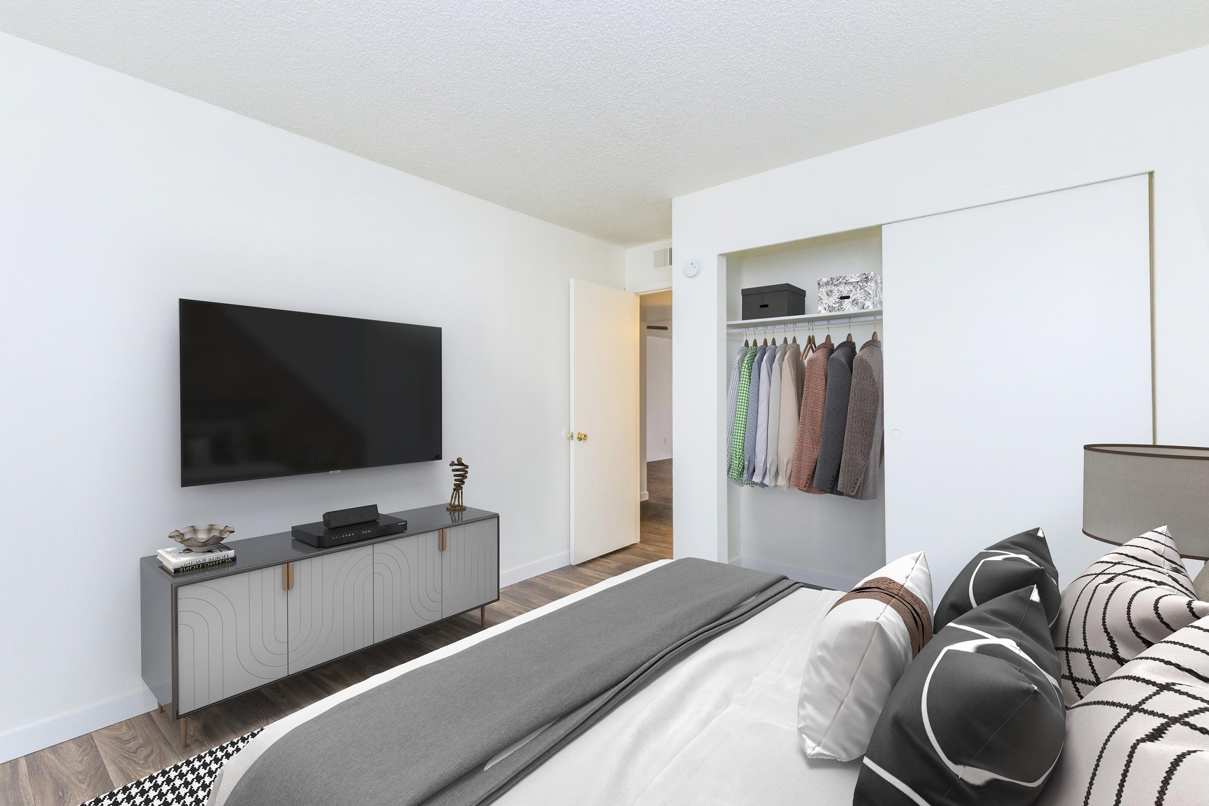 A modern bedroom featuring a large flat-screen TV mounted on the wall, a neatly made bed with decorative pillows, and a stylish dresser. In the wardrobe, neatly hung clothes are visible, and the room has light-colored walls and wooden flooring, creating a bright and inviting atmosphere.