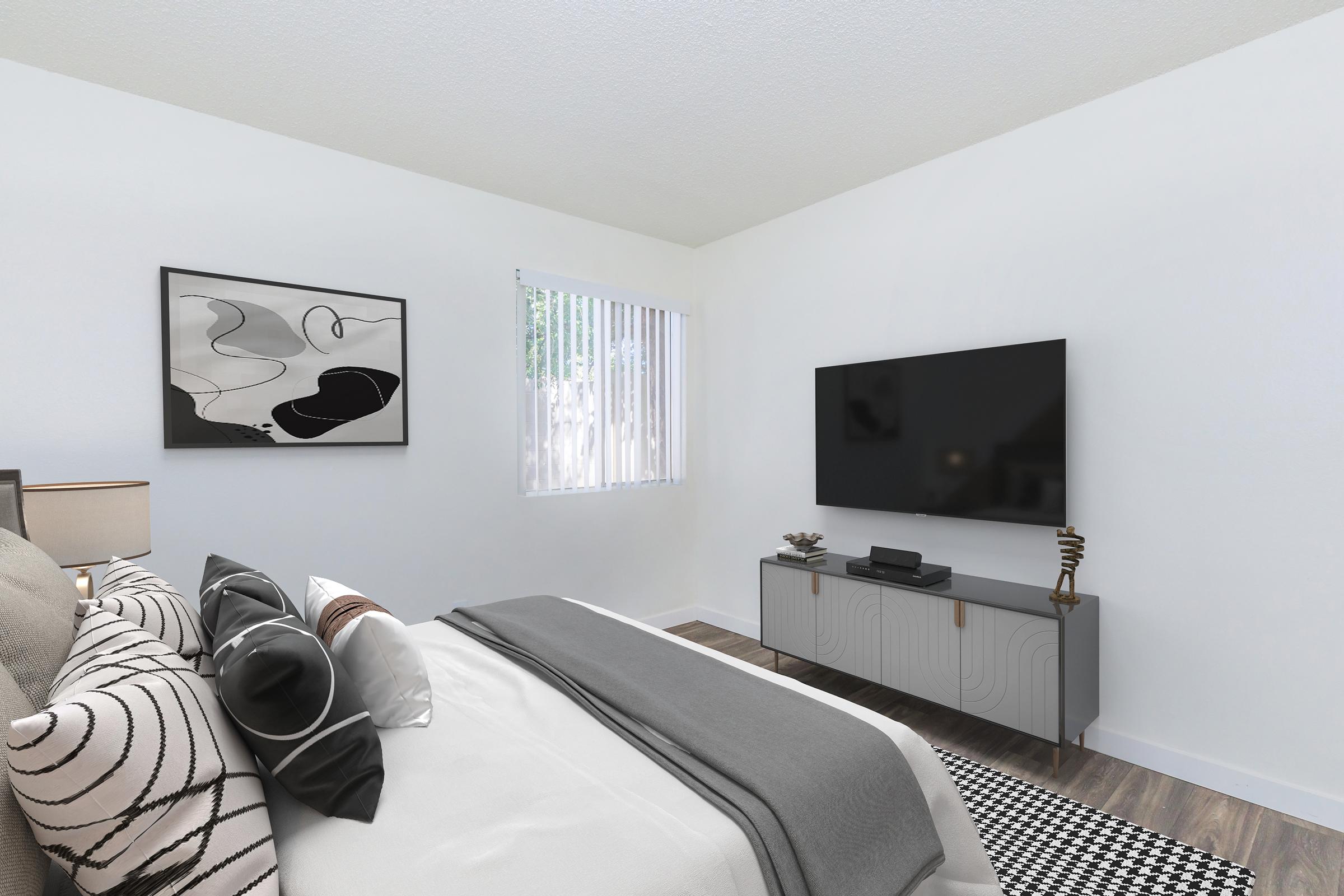 A modern bedroom featuring a large bed with decorative pillows, a sleek gray TV stand with a flat-screen TV, and a piece of abstract art on the wall. Natural light filters through window blinds, illuminating the minimalistic design with a neutral color palette. A patterned rug enhances the space.
